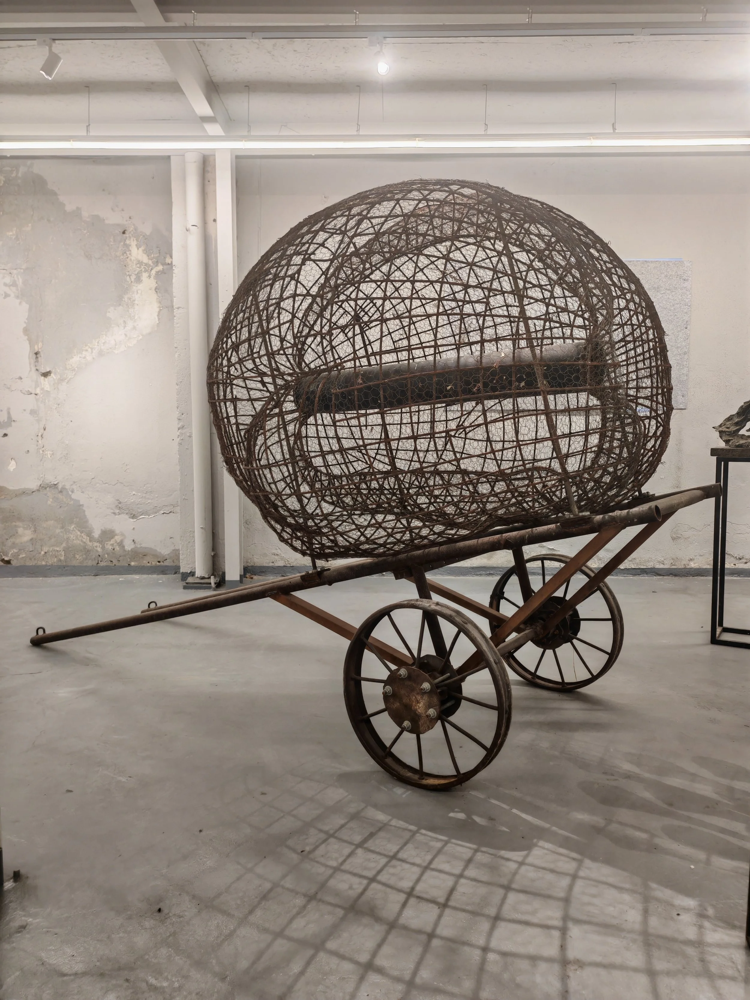 An old, rusted wooden cart with a large spherical wireframe sculpture resting inside it, in an industrial-style gallery space with white walls and concrete flooring.