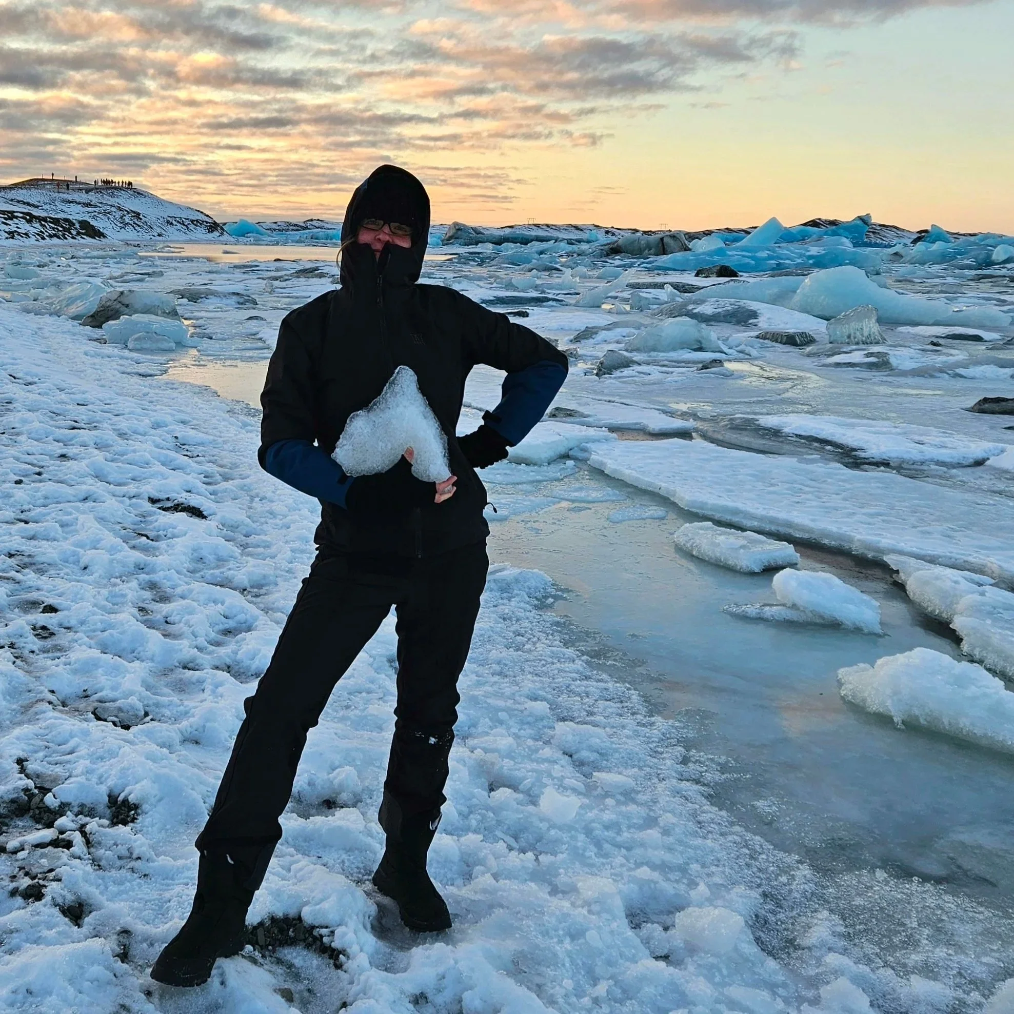 Person dressed in winter clothing holding a piece of ice on snow-covered landscape with ice chunks and a snowy hill in distance during sunset or sunrise.