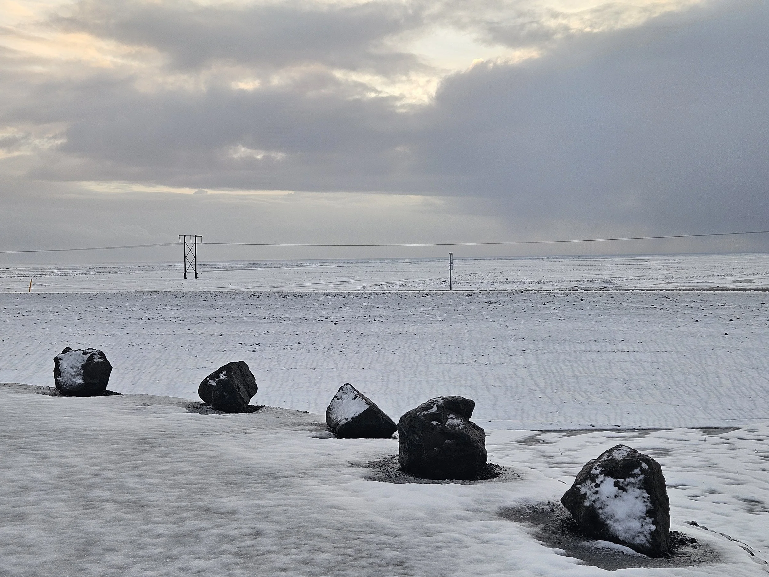 A snow-covered landscape with large rocks in the foreground, a power line running across the scene, and a cloudy sky overhead.