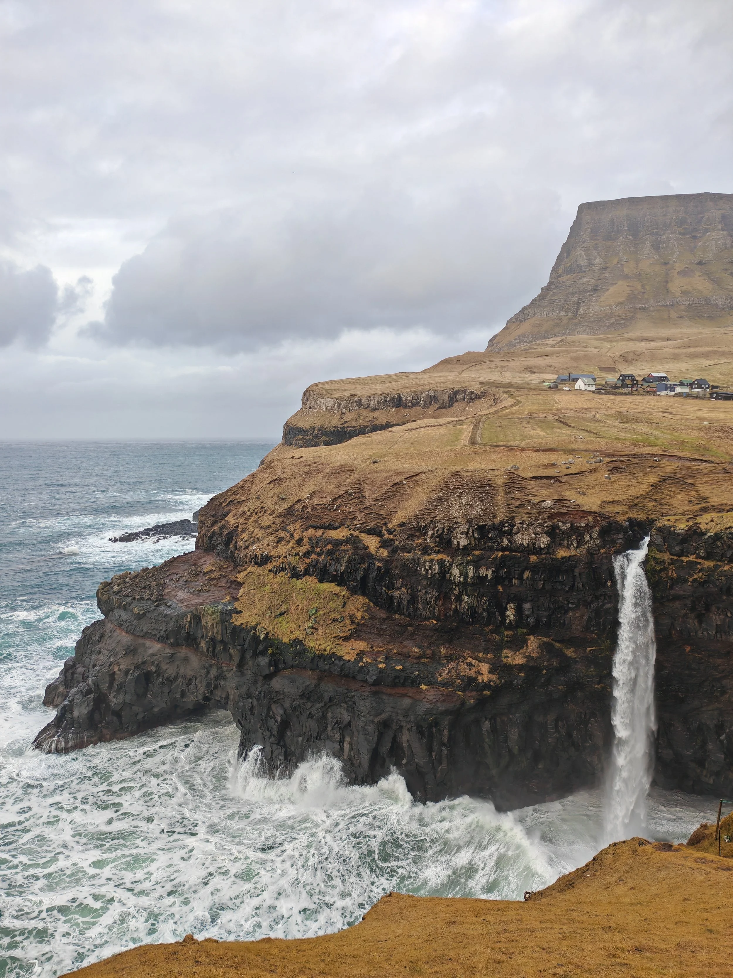 Cliffs overlooking the ocean with a waterfall cascading down from the edge, and a small village with houses near the base of the cliffs under a cloudy sky.