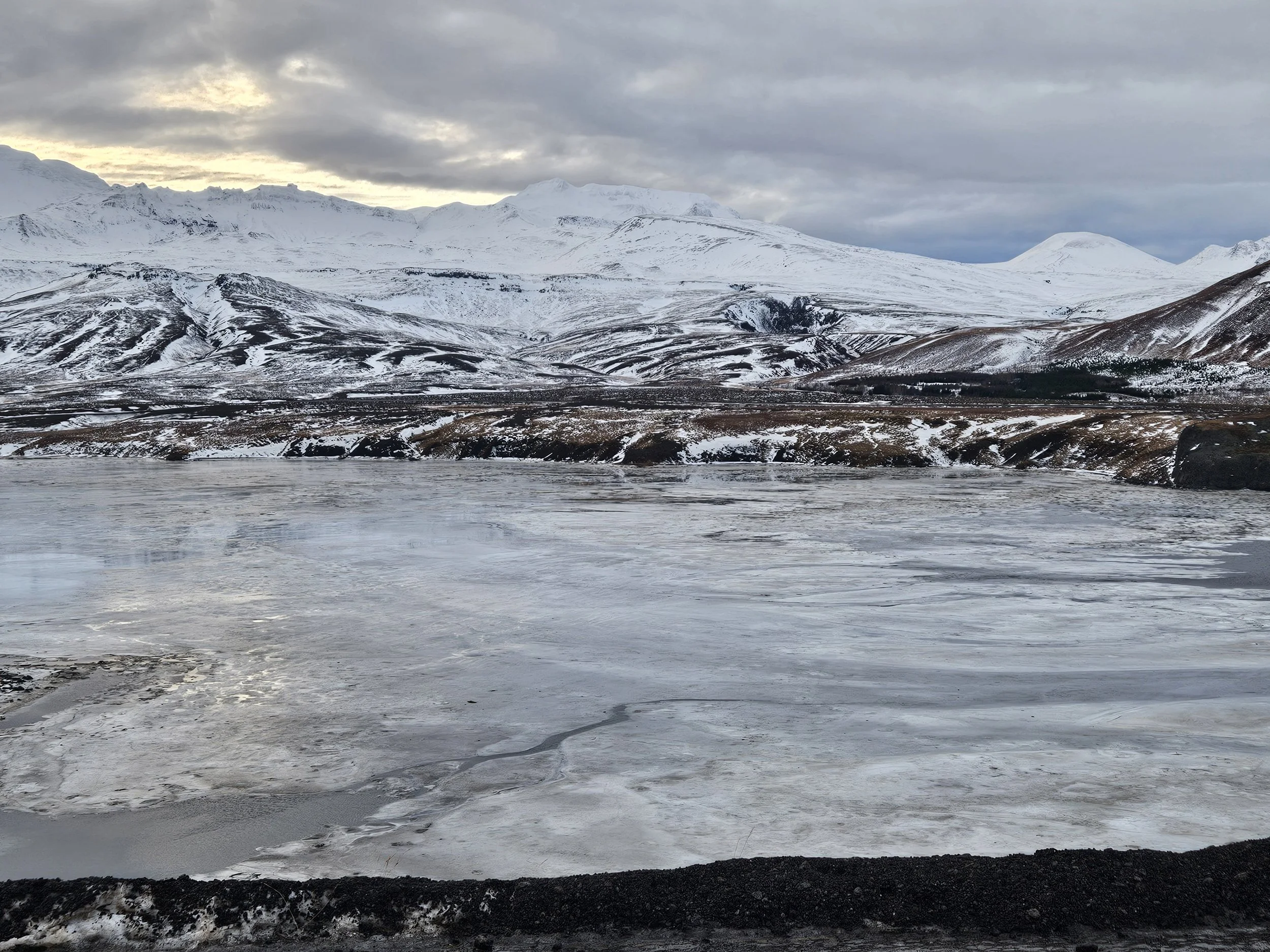 Snow-covered mountains with partly cloudy sky, frozen body of water in the foreground, and patches of dark earth on the shoreline.