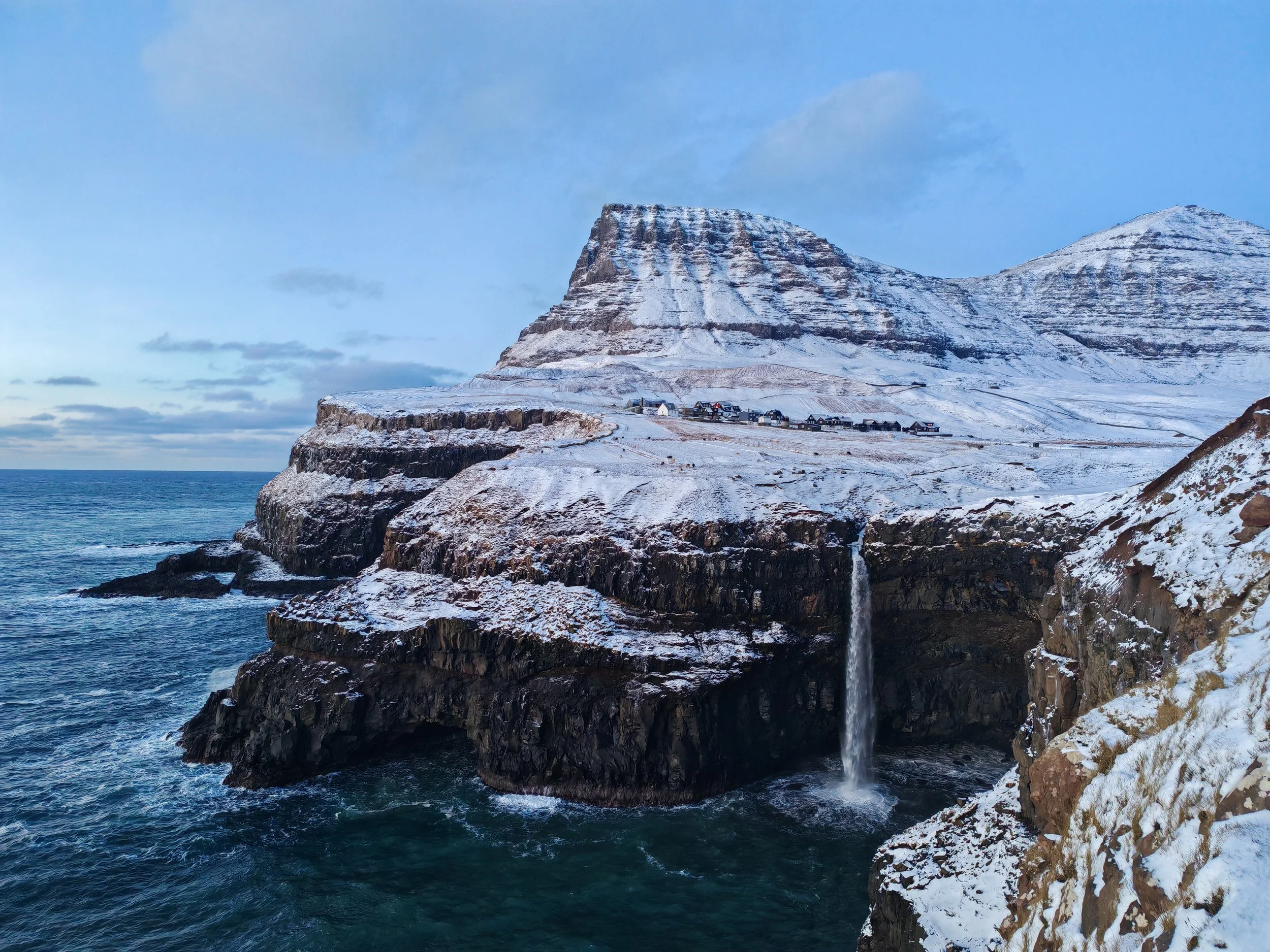 Snow-covered cliffs overlooking the ocean with a small village at the base, under a partly cloudy sky.