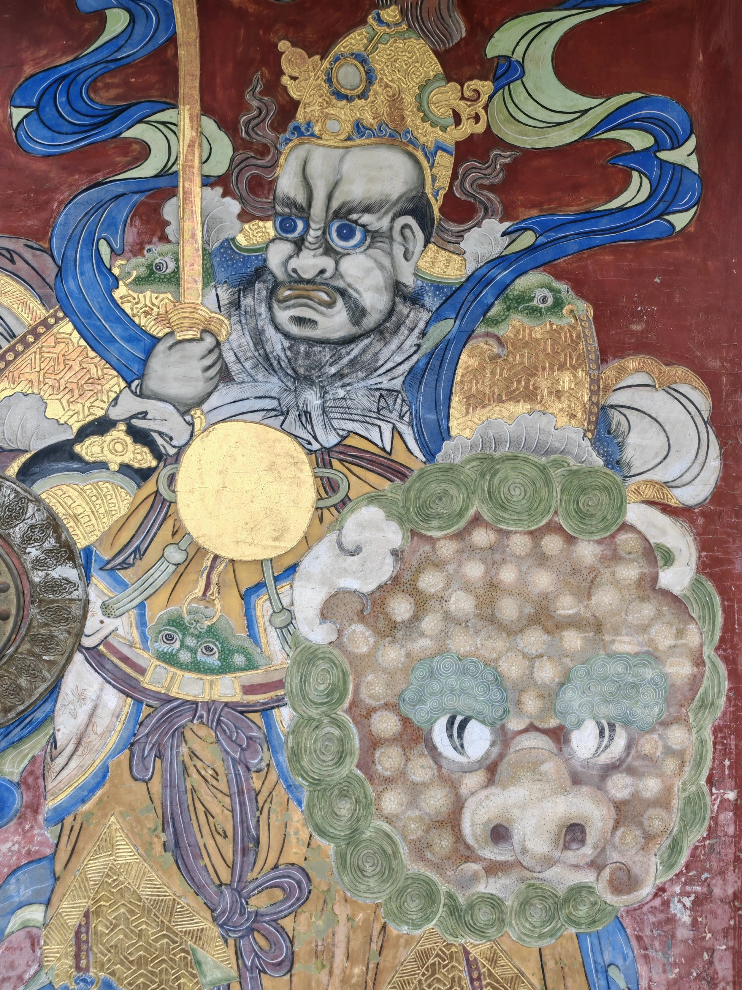 Detail of a traditional Asian mural featuring a fierce warrior with blue eyes, ornate armor in gold and green, and a headdress. The warrior is holding a sword and surrounded by stylized clouds and swirling patterns.