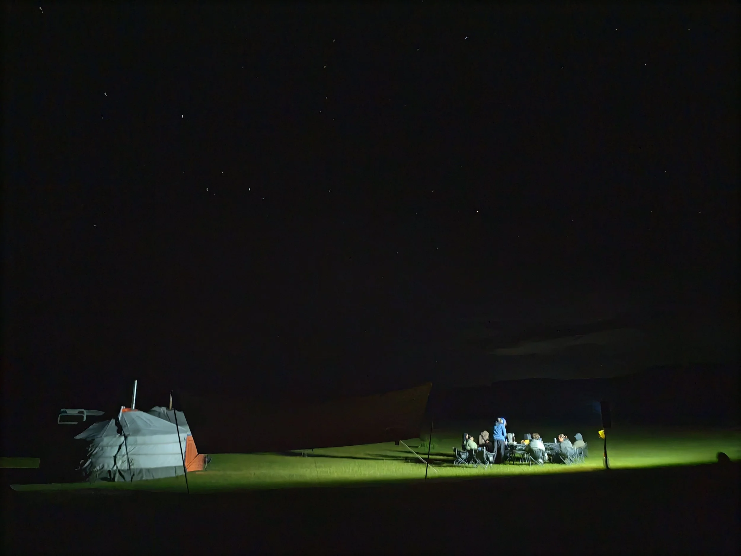 Nighttime camping scene with tents and people sitting around illuminated area, dark sky with stars visible.