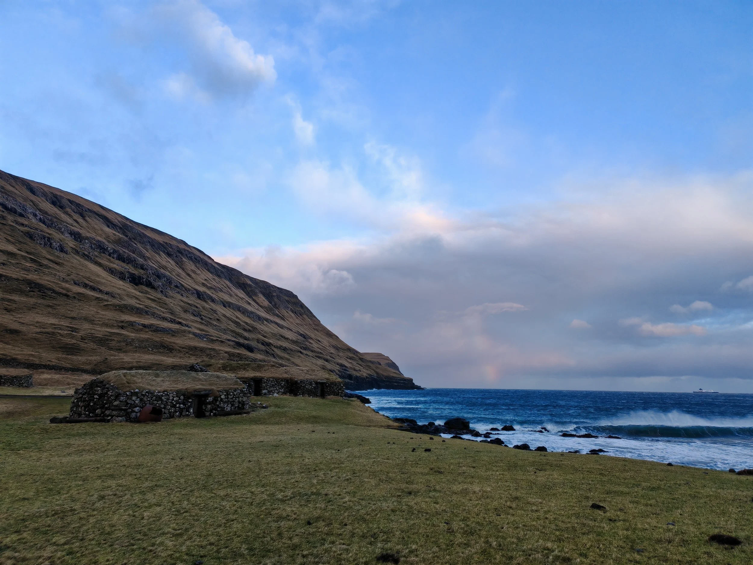 A coastal landscape with a grassy foreground, stone structures, a rocky shoreline, a blue ocean, a mountain slope on the left, and a sky with clouds overhead.