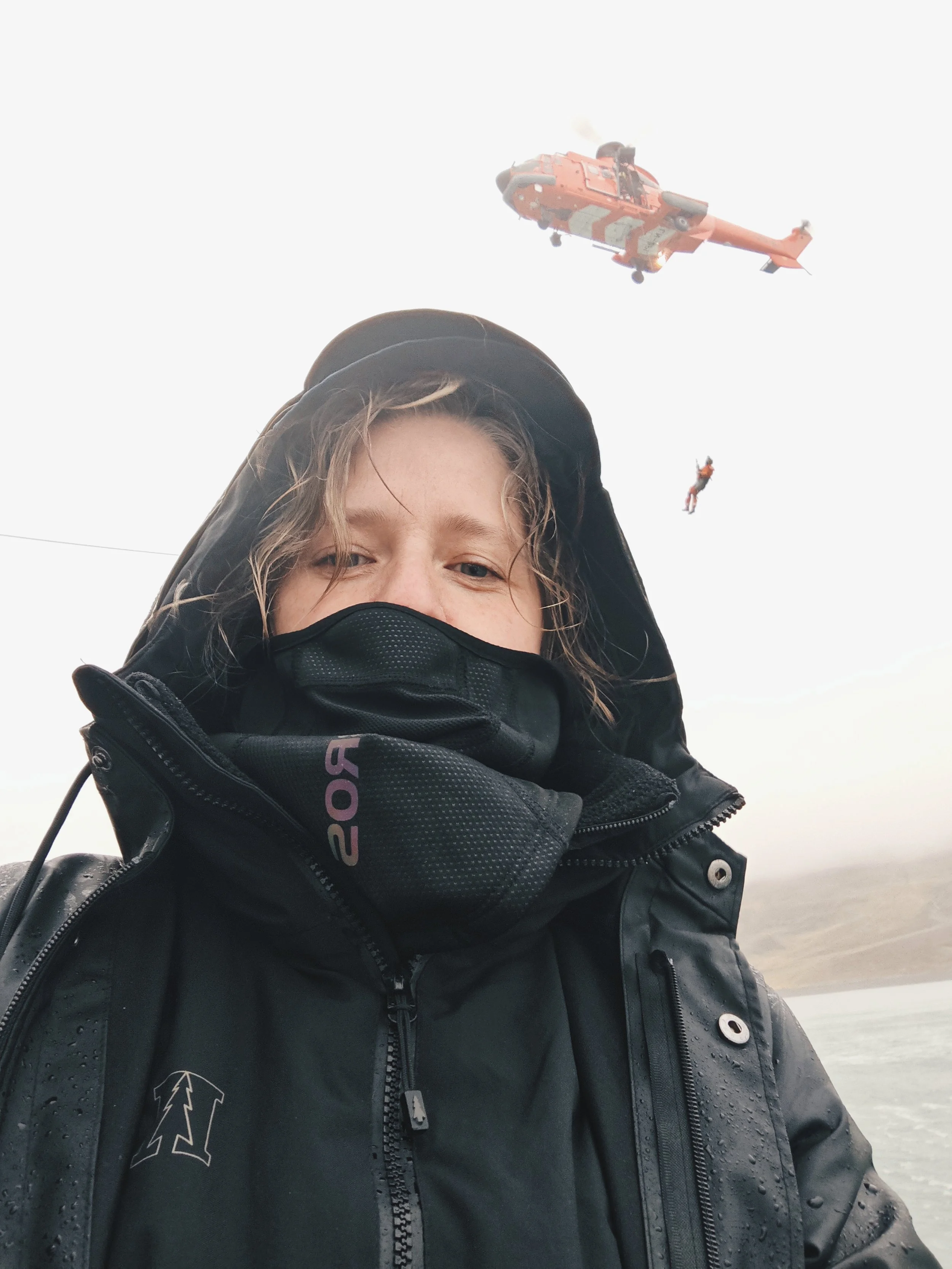 A person wearing a black jacket, black face mask, and a hat, standing outdoors near water, with a helicopter and a person parachuting in the background.