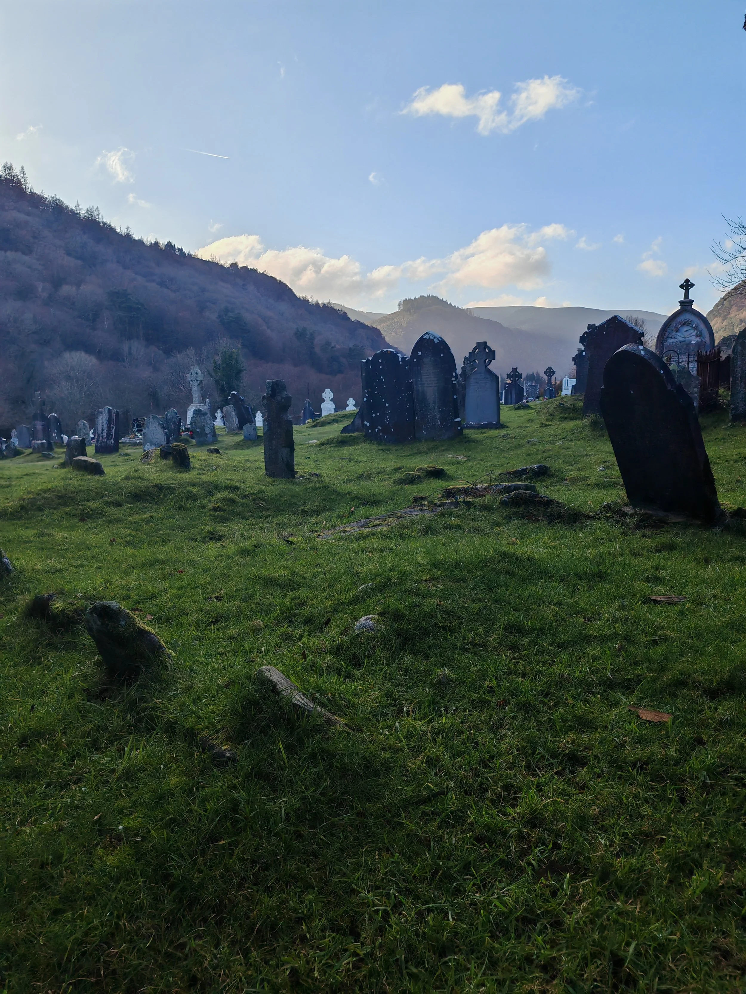 A cemetery with numerous tombstones and crosses on green grass, surrounded by mountains and trees, under a blue sky with clouds.