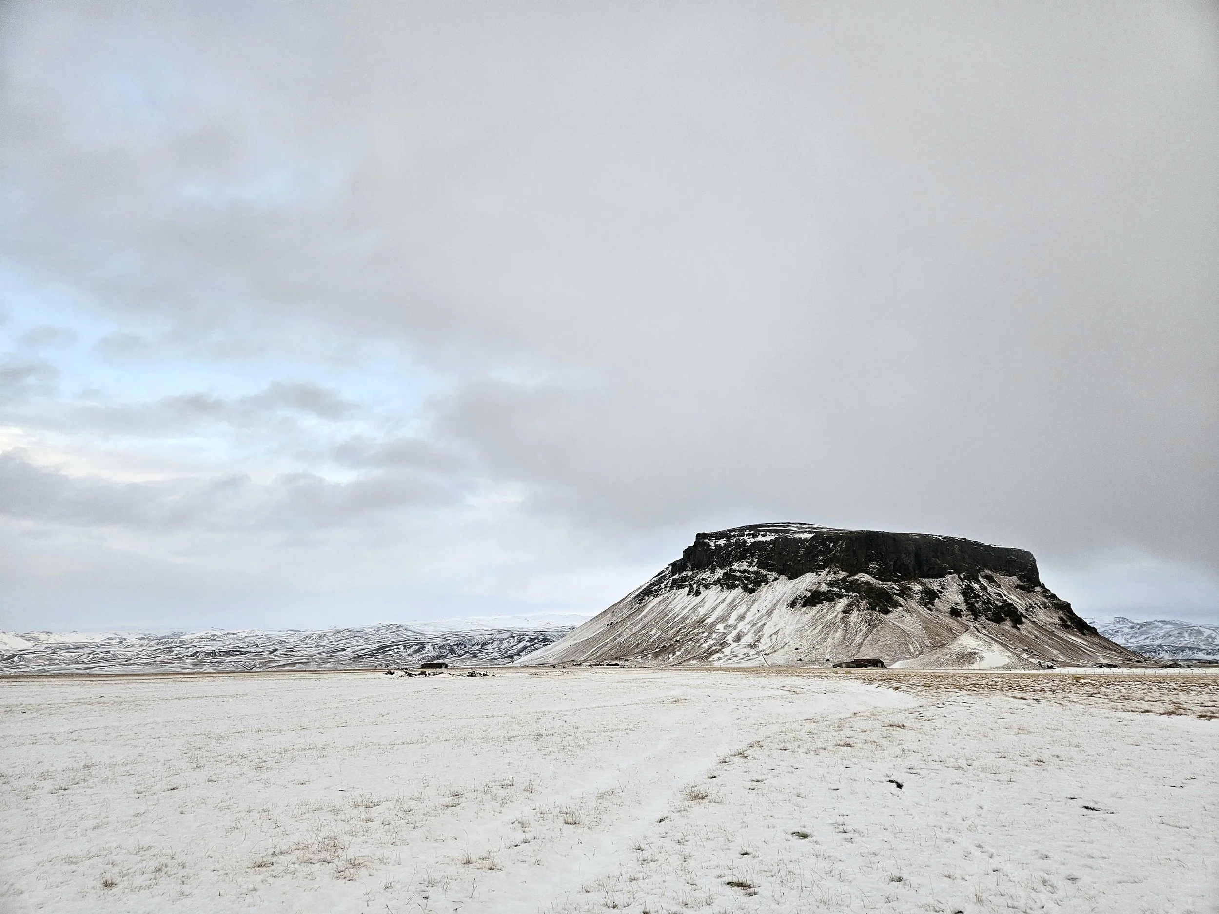 Snow-covered plain with a large, flat-topped mountain or mesa with snowy slopes in the distance under a cloudy sky.