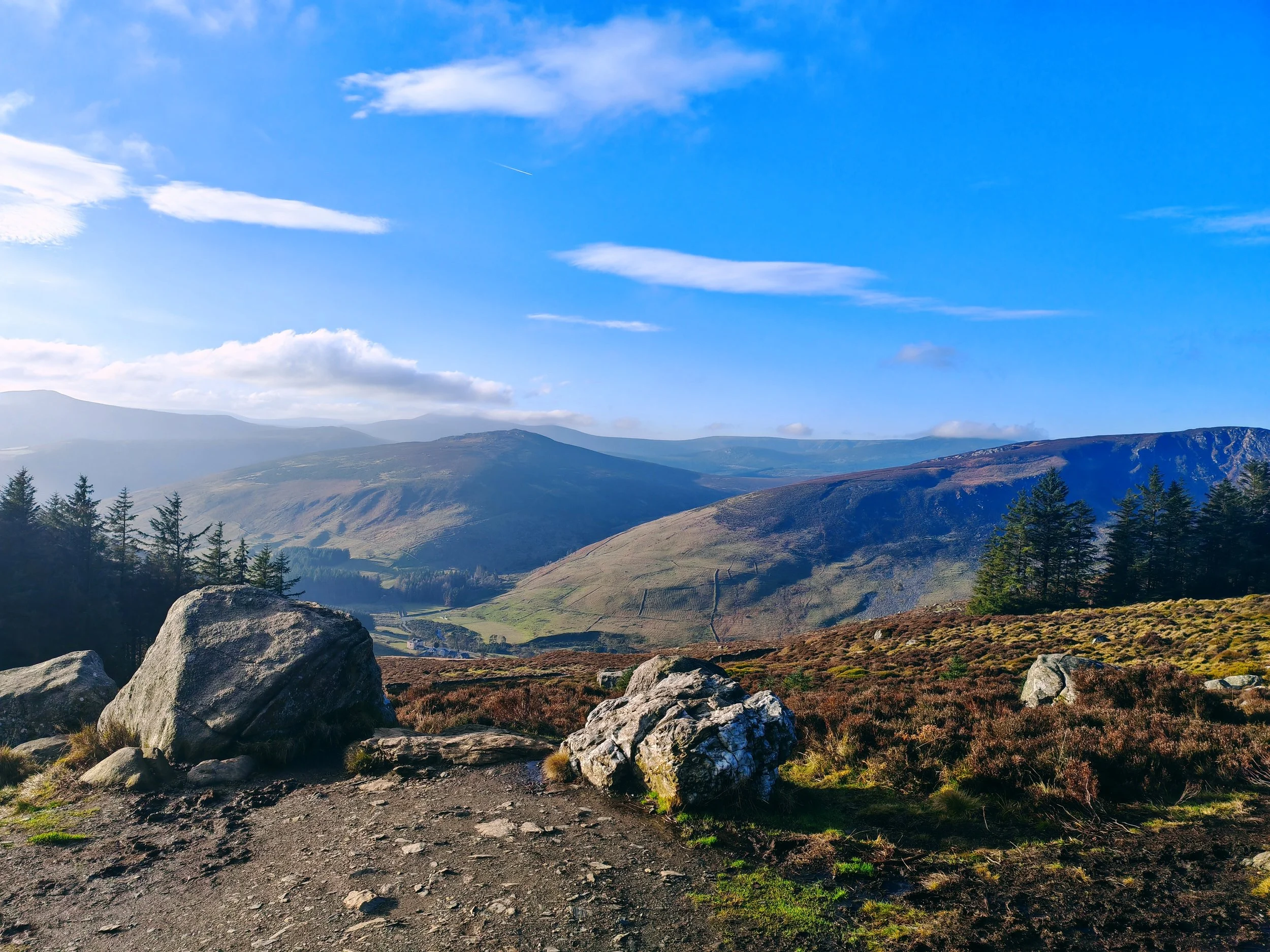 Scenic mountain landscape with large rocks in the foreground, green trees, rolling hills, and distant mountains under a partly cloudy blue sky.