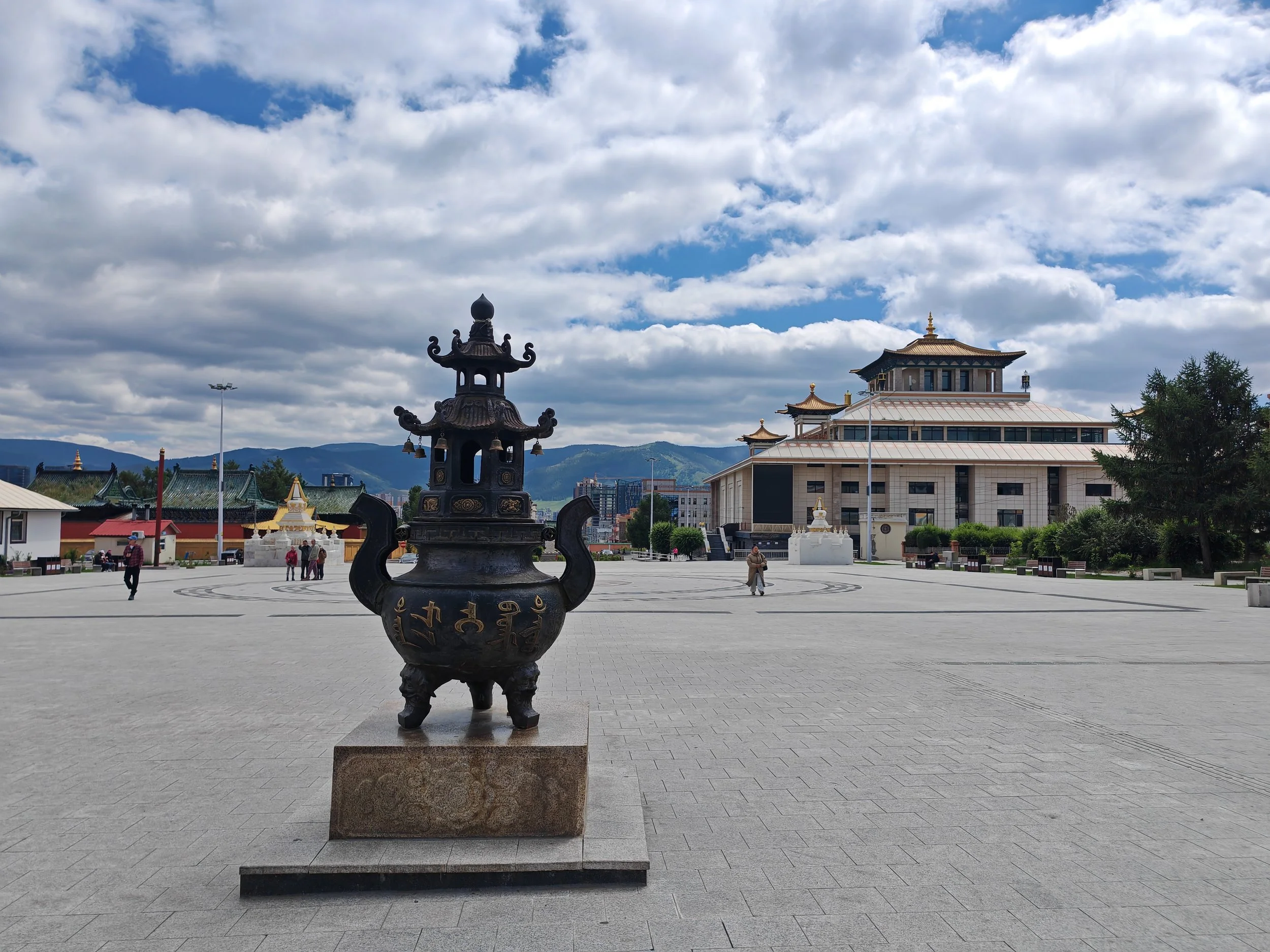 A large open plaza with a traditional black metallic statue in the foreground, featuring multiple tiers with decorative elements. In the background, there is a multi-story building with traditional Asian architectural design, including a curved roof 