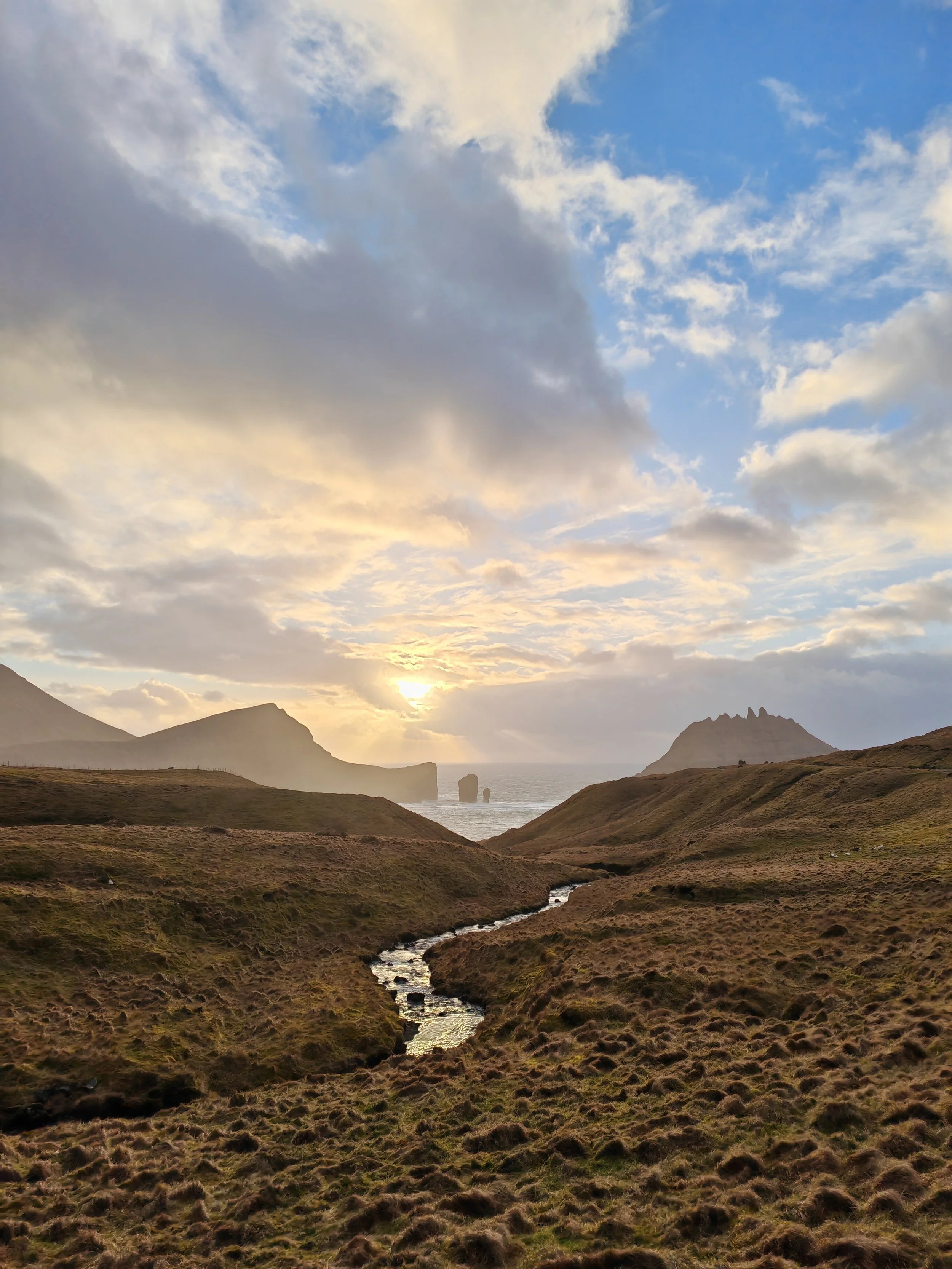 A rugged landscape with rolling hills, a small winding stream, distant rocky cliffs, and a setting sun shining through scattered clouds in the sky.