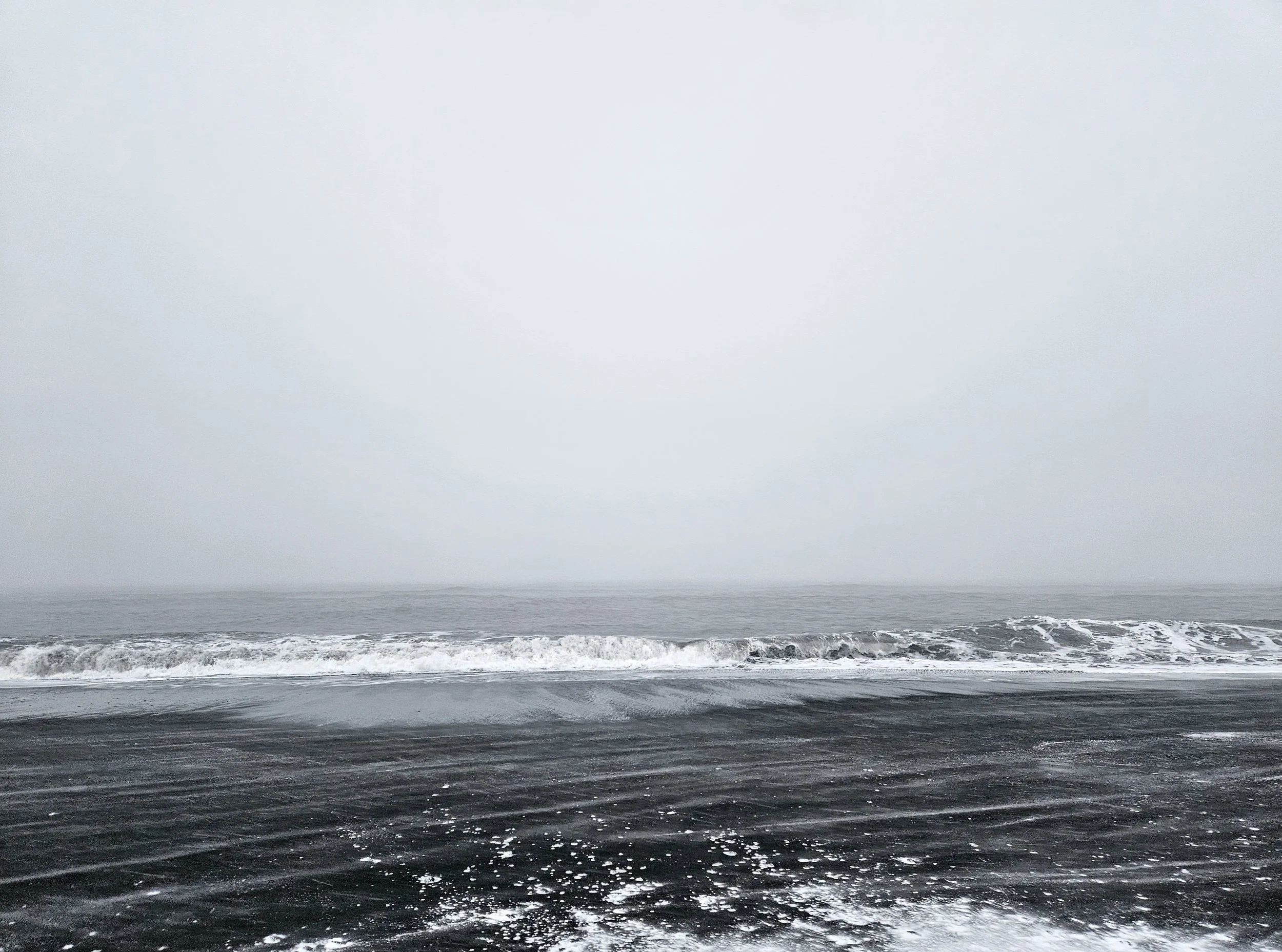 Overcast sky with ocean waves crashing on a dark sandy beach.