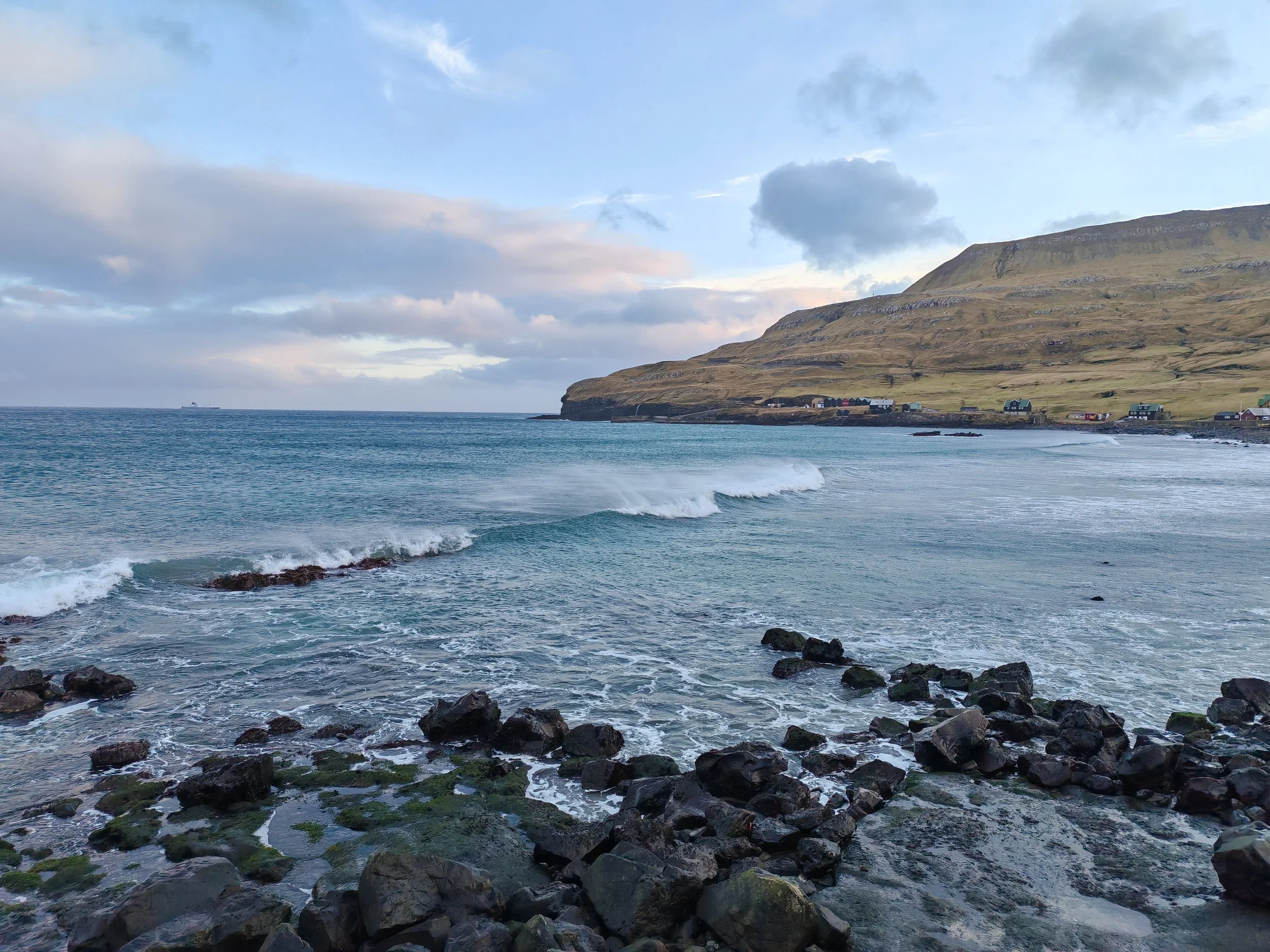 A coastal scene with rocky shoreline in the foreground, gentle waves, a blue ocean, a hillside with sparse houses, and a partly cloudy sky.