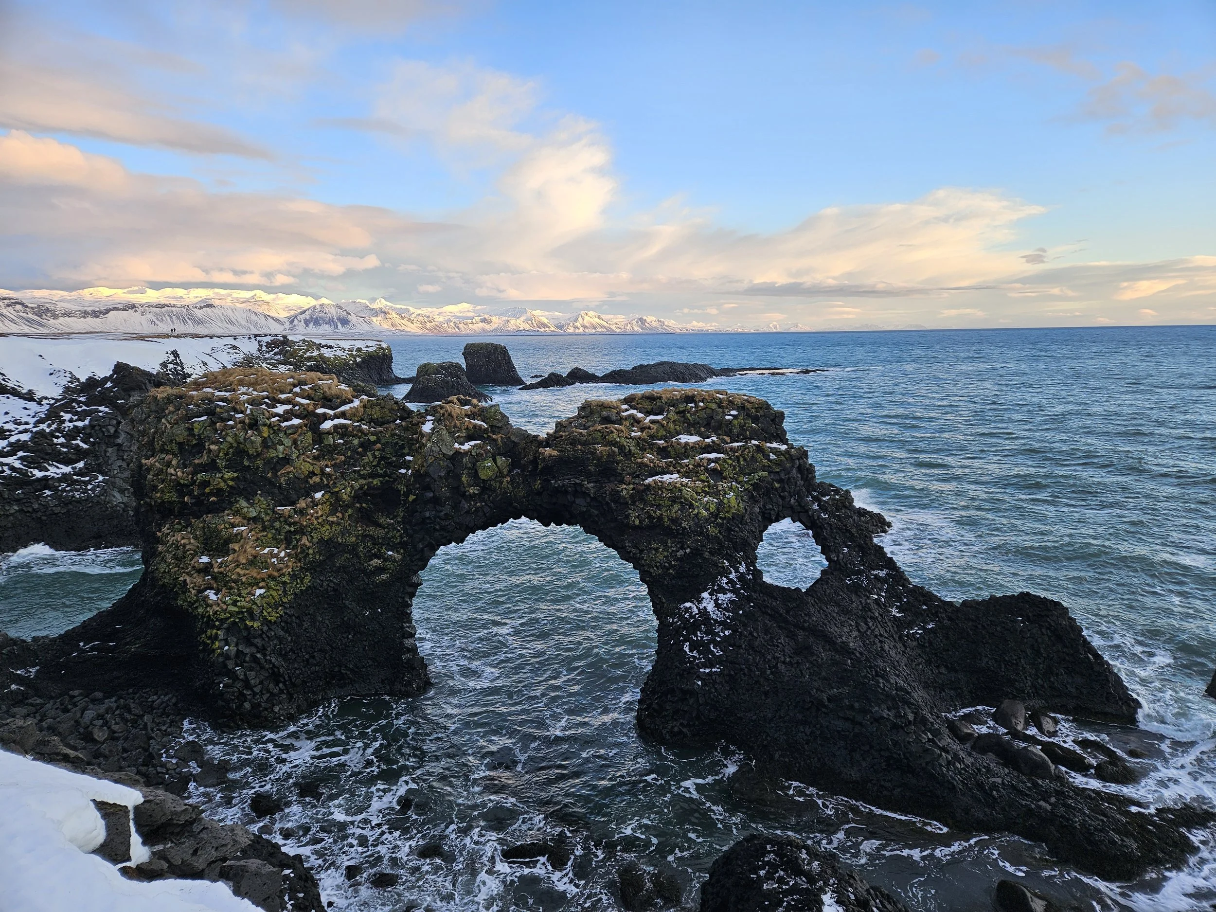A rocky, snow-dusted coastline with black volcanic rocks in the foreground, dark blue ocean waves, snow-capped mountains in the background, and a partly cloudy sky.