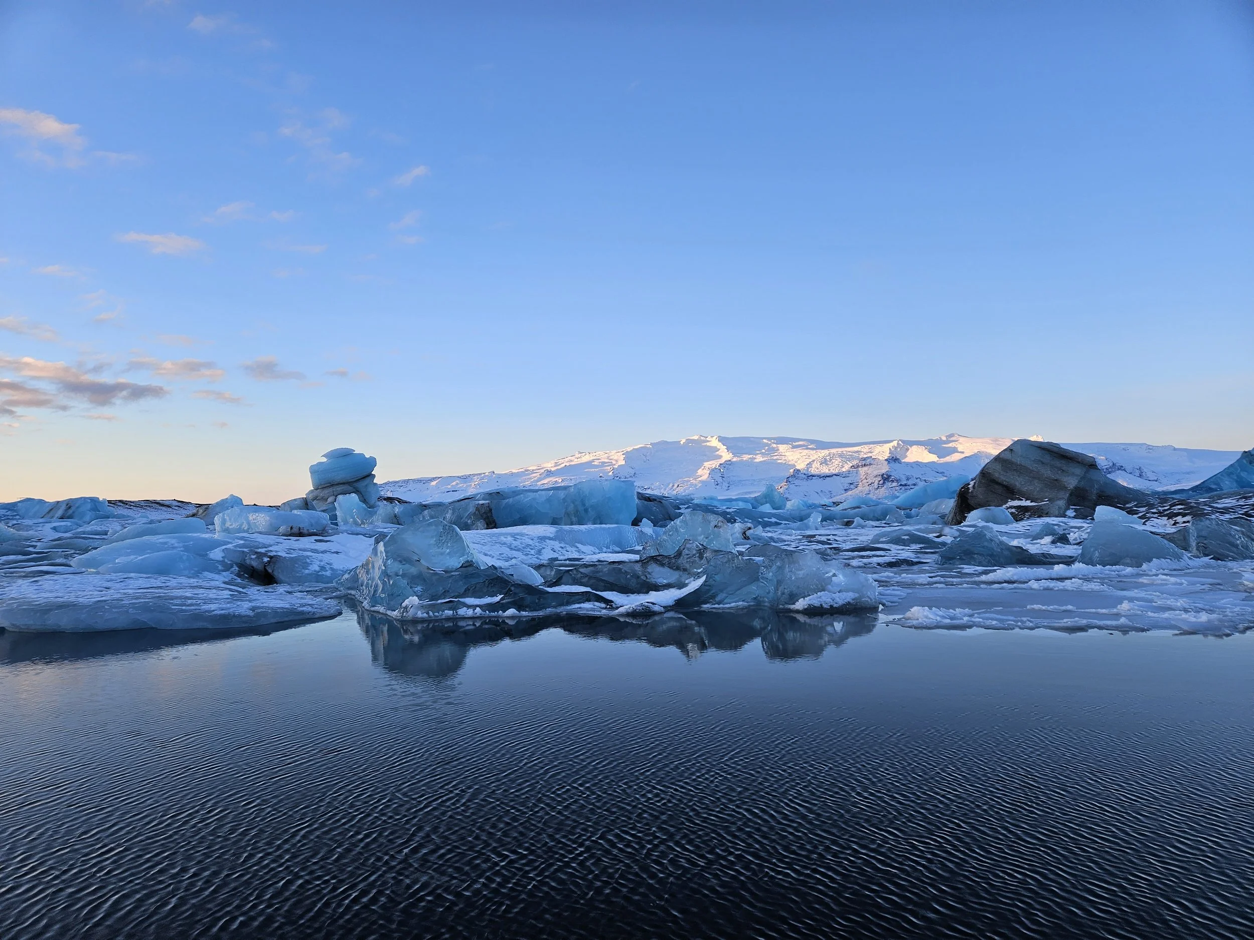 Frozen landscape with glaciers, snow-covered mountains in the background, and a calm body of water reflecting the sky.