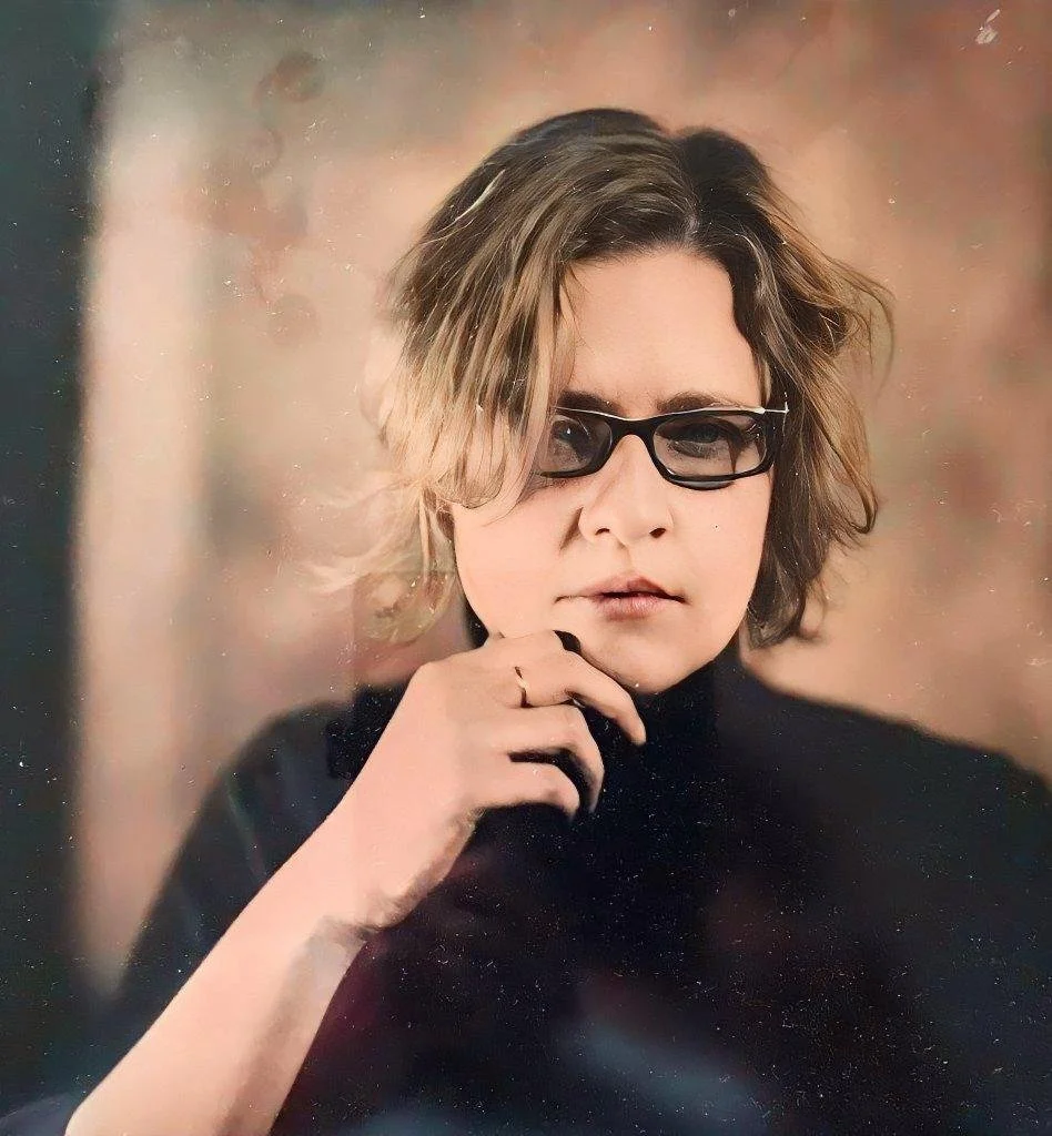 Portrait of a woman with short, wavy hair wearing glasses and a black turtleneck, with her hand near her chin, looking serious.