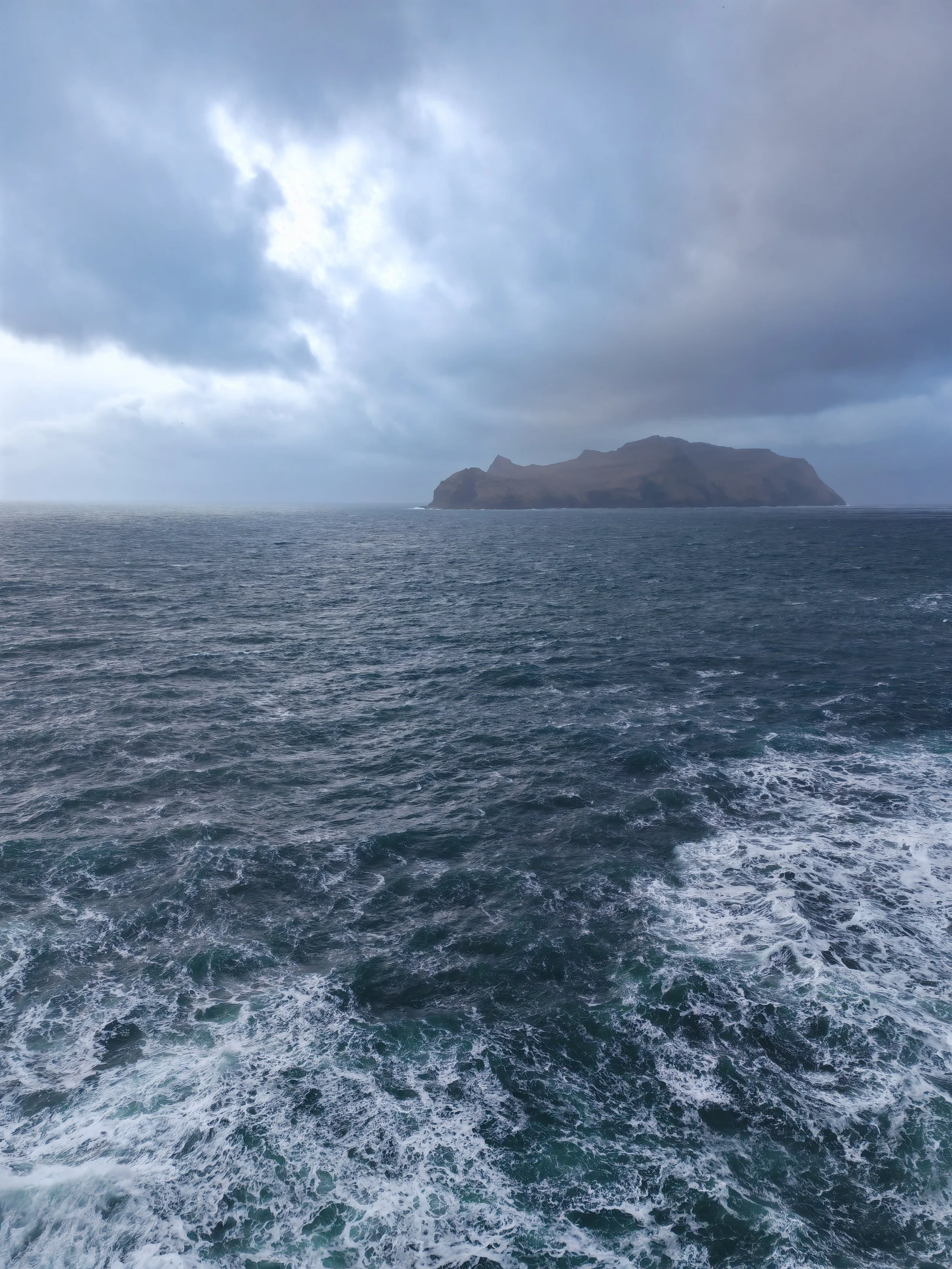 View of a large island with steep cliffs and hills, surrounded by ocean with visible waves and water turbulence. The sky is cloudy and overcast.