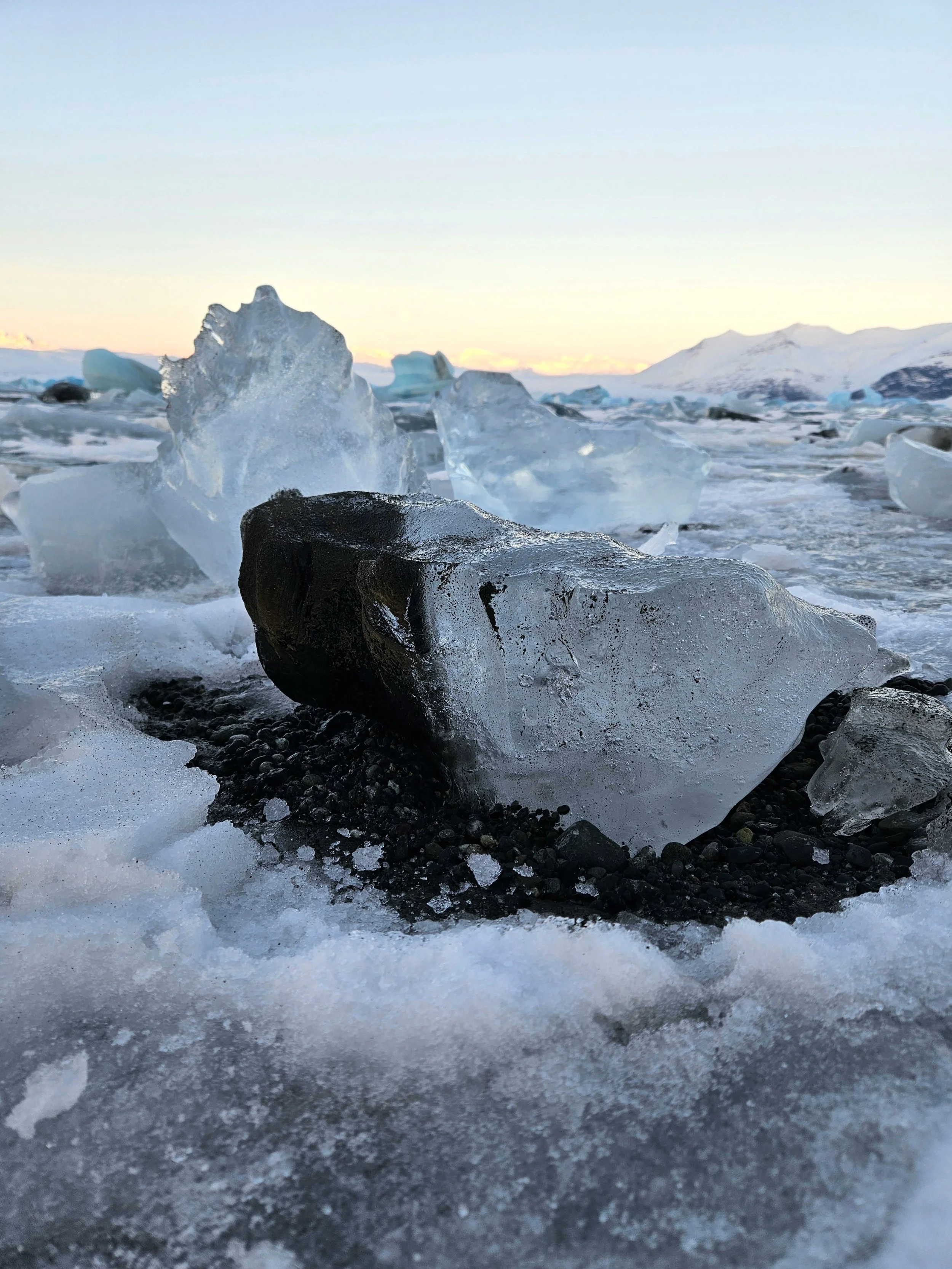 Ice chunks and rocks on a snowy landscape with mountain in the background and sunrise or sunset sky.