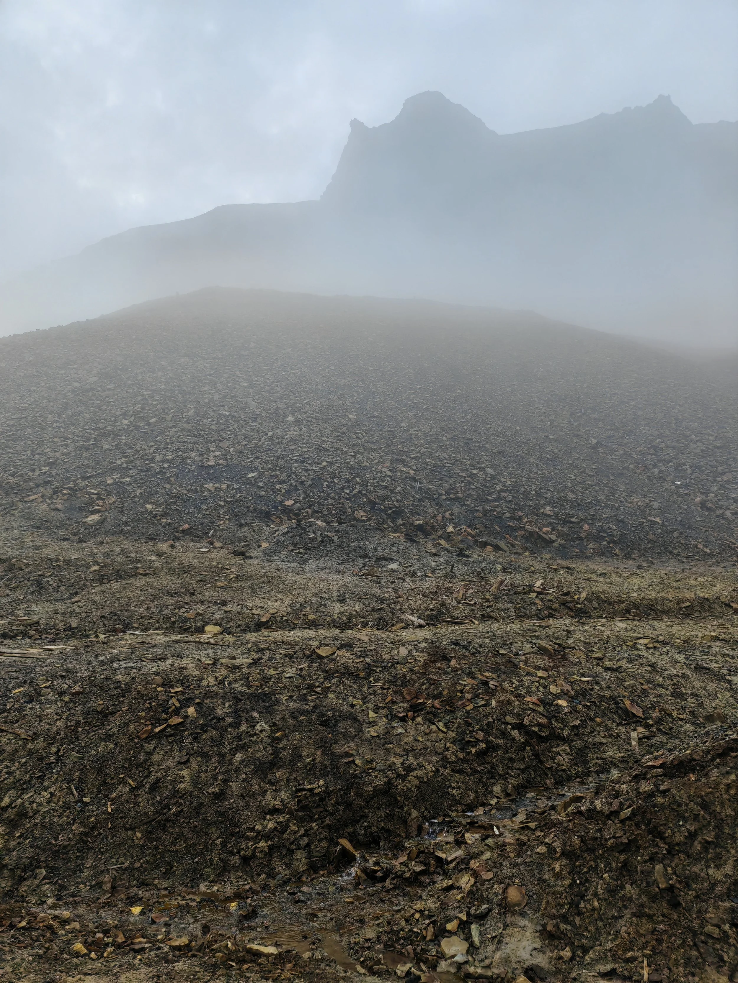 A foggy mountain landscape with a large rocky slope in the foreground and a mountain peak in the background, partially obscured by mist.