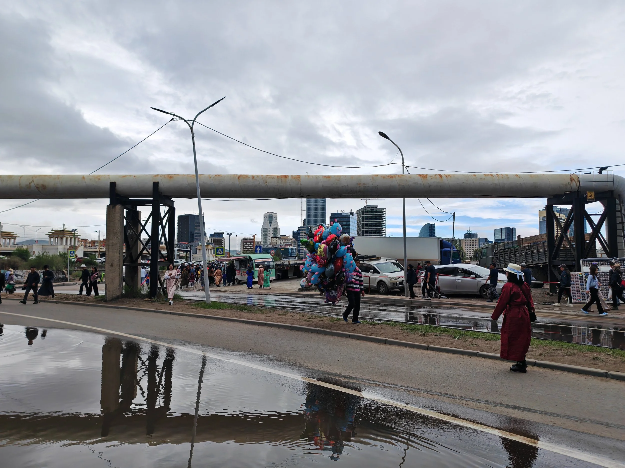 Urban scene with people walking along a street, colorful balloons for sale, and a large overhead pipe or pipeline with city buildings in the background under cloudy sky.