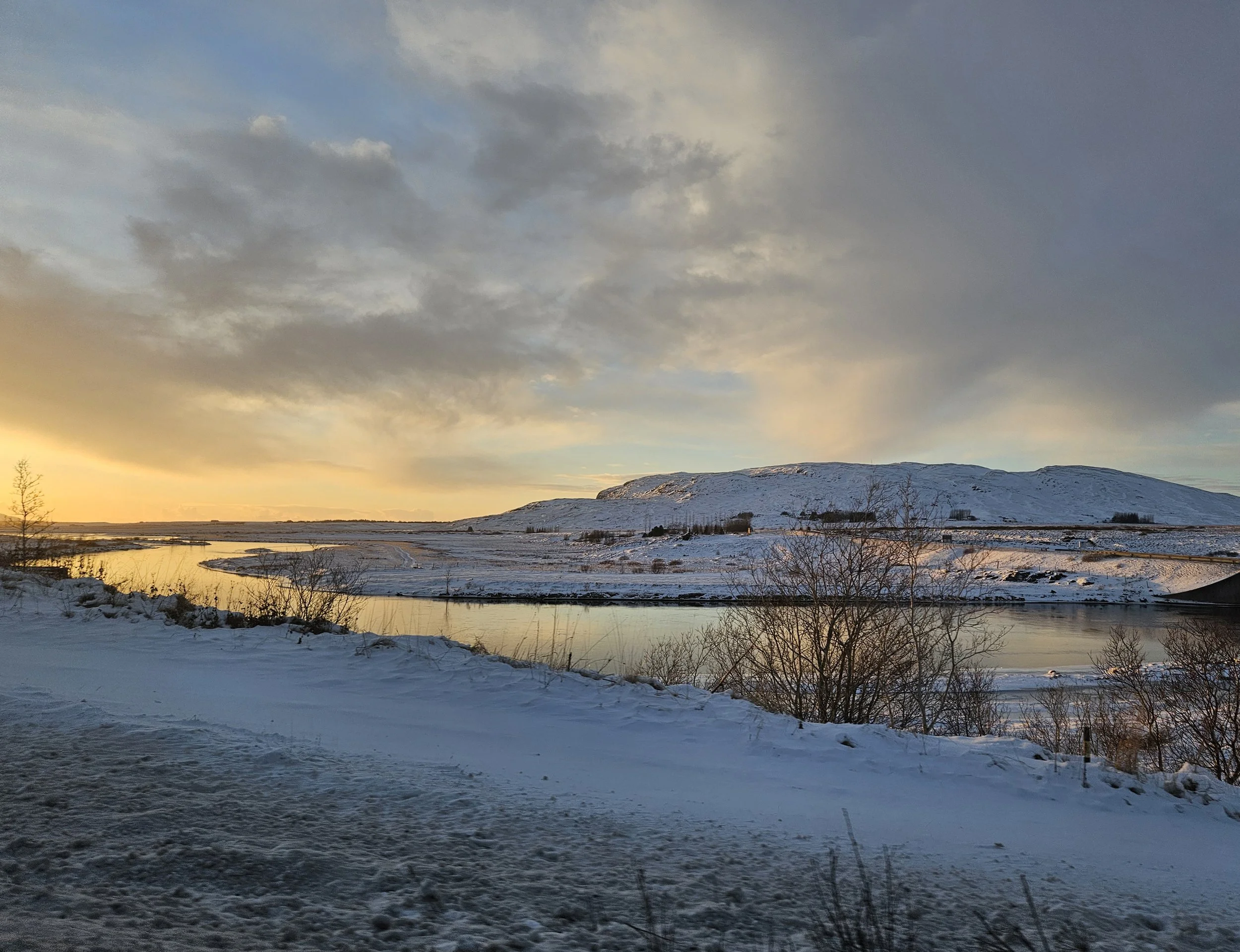 Snow-covered landscape with a river, hills, and a cloudy sky at sunset.