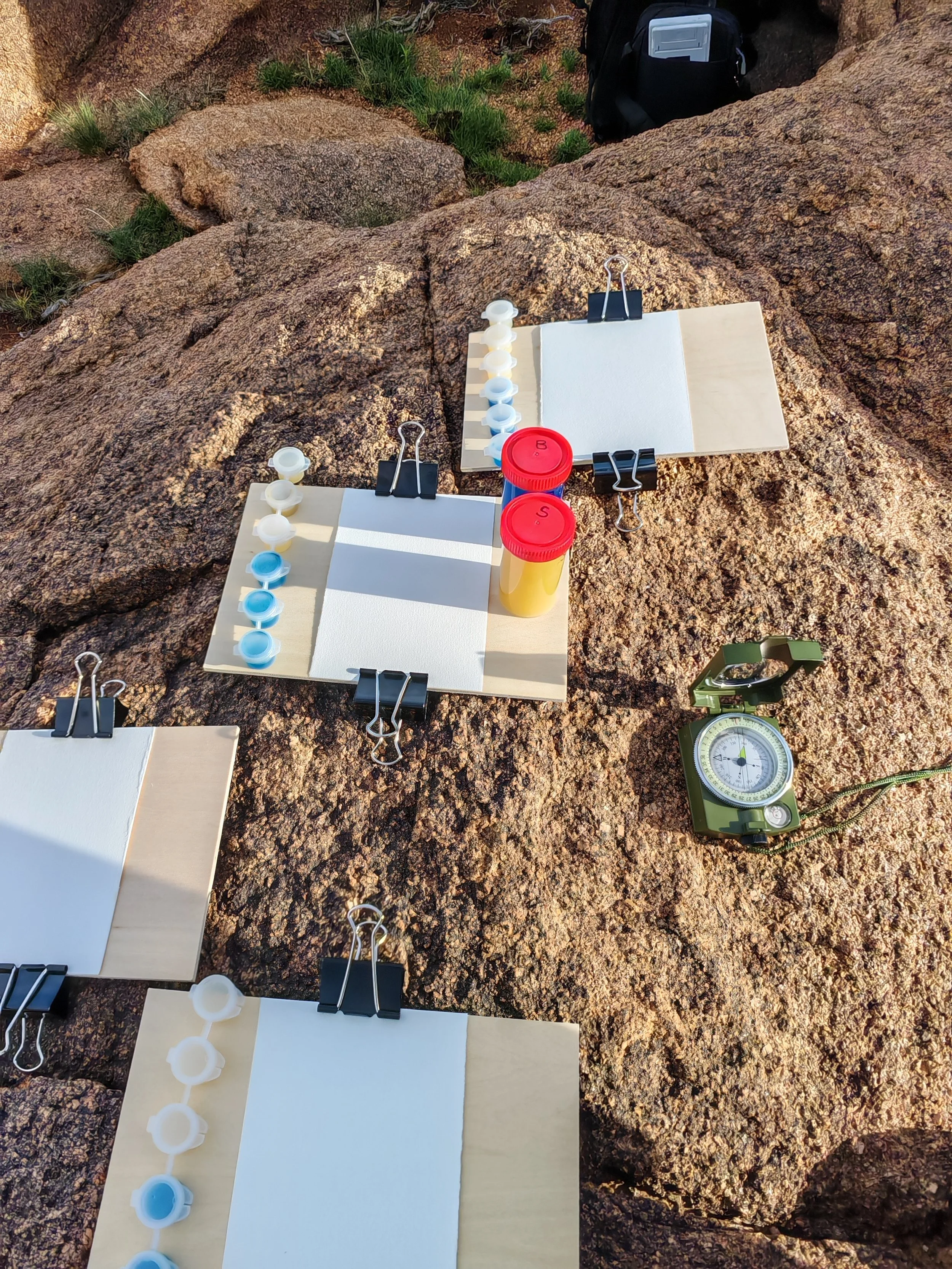Medical supplies for field testing, including paper, gloves, and small bottles, arranged on rocks outdoors with a black backpack and a green compass nearby.