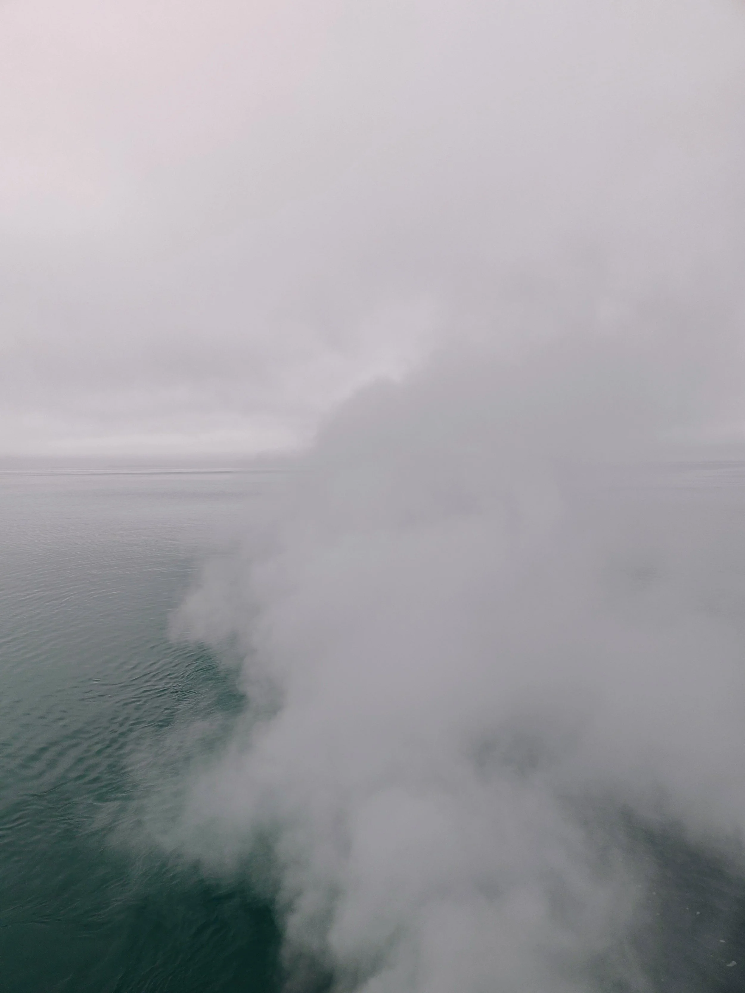 View of ocean water with a large plume of steam or smoke rising from underwater, surrounded by gray clouds and overcast sky.