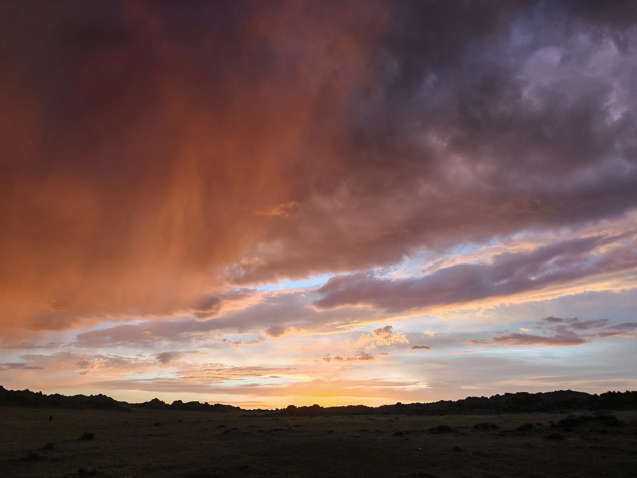 A landscape view of a sunset with dark clouds in the sky, illuminating the horizon with orange and yellow hues, and a silhouette of trees and hills in the foreground.