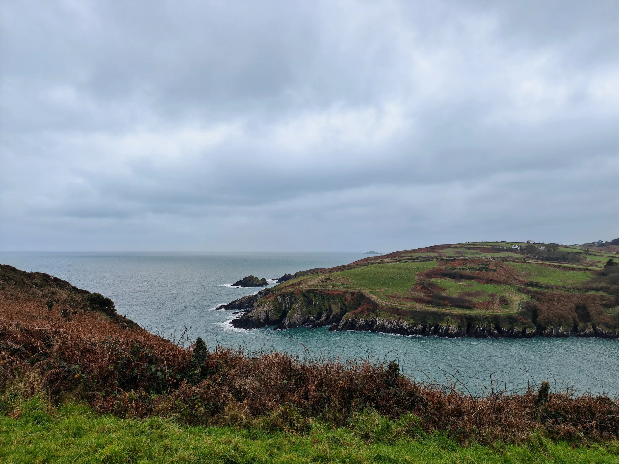 Cliffs along a coastline under a cloudy sky with patches of green fields and a distant small house on the hill.