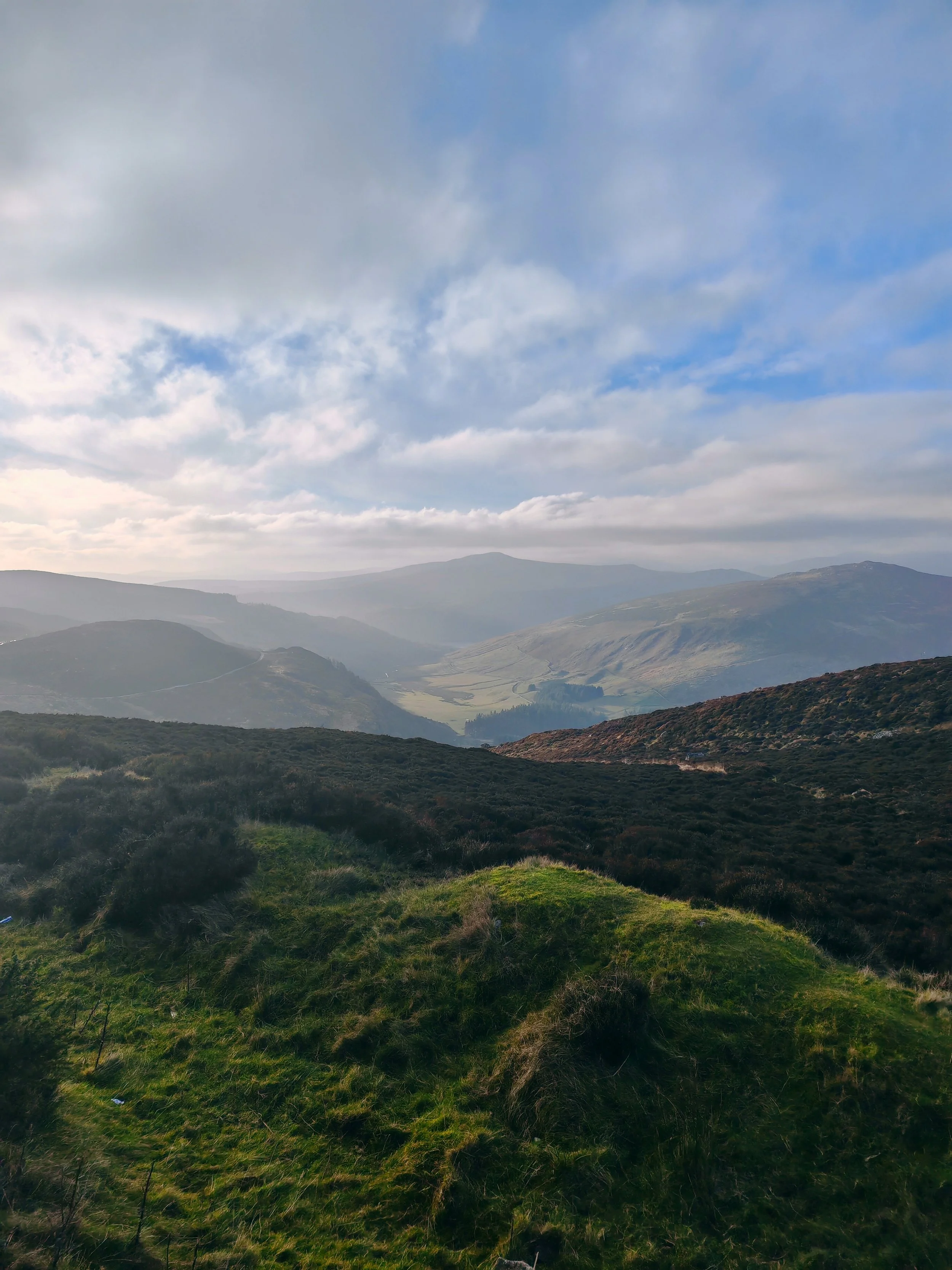 A landscape of rolling green hills, distant mountains, and a cloudy sky with blue patches.