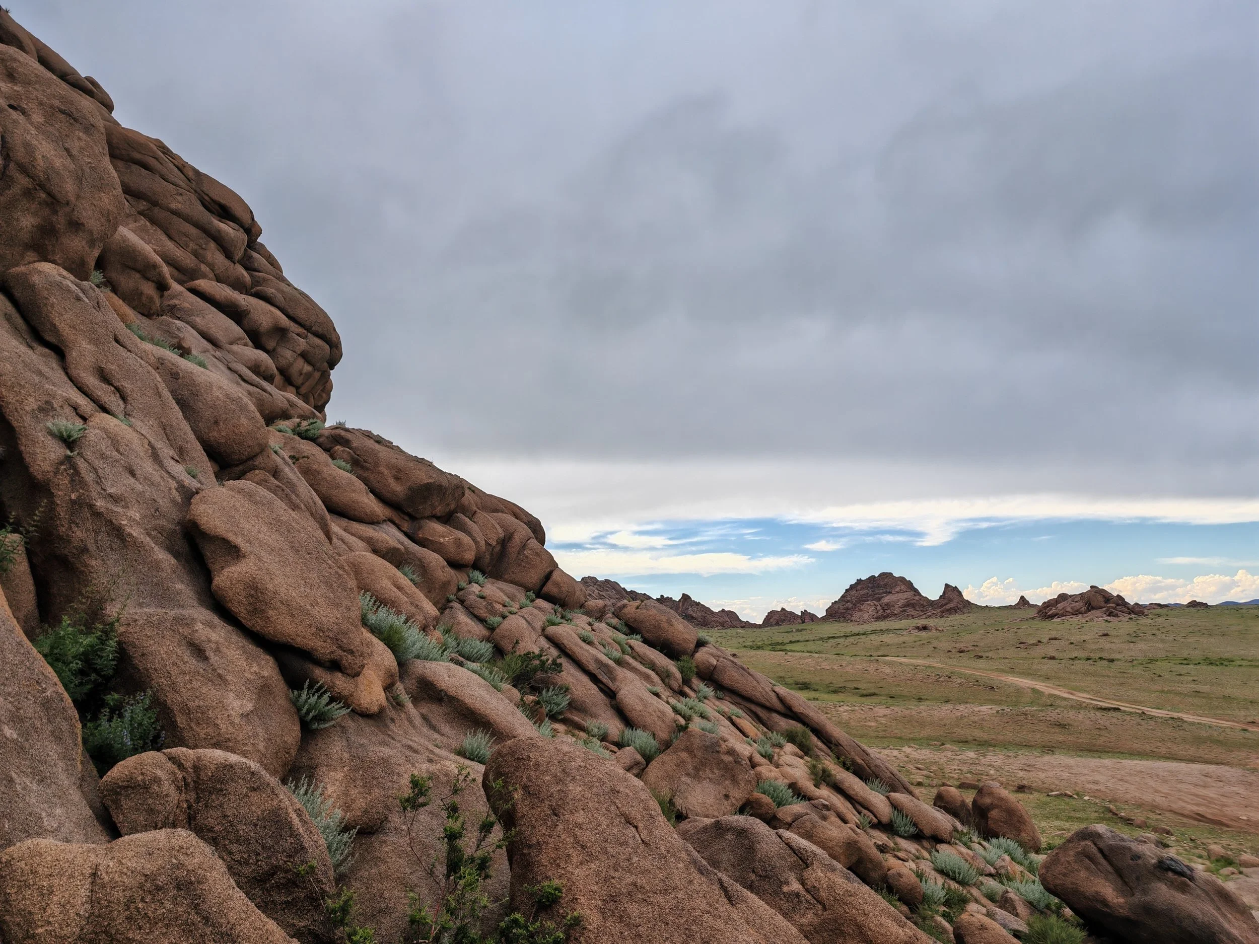 A landscape with large red rocks on the left and open grasslands with small rocks and shrubs extending toward the horizon under a cloudy sky.