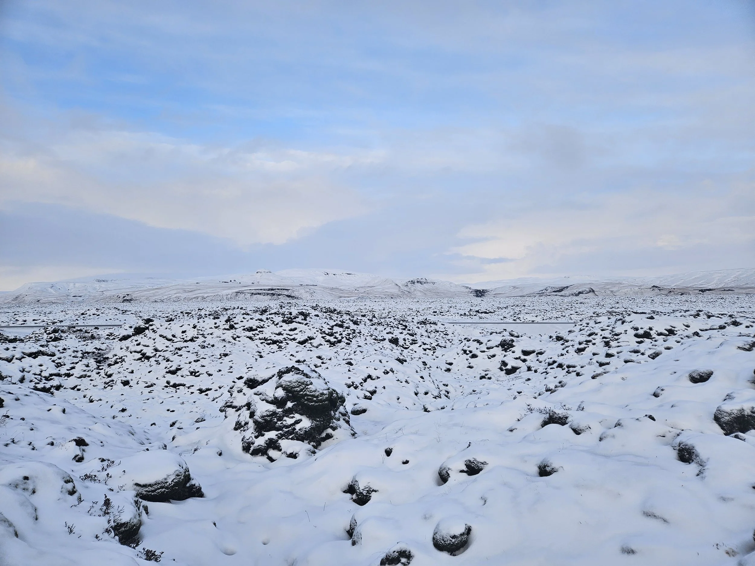 Snow-covered landscape with dark rocks scattered across the ground, and distant snow-covered hills under a partly cloudy sky.