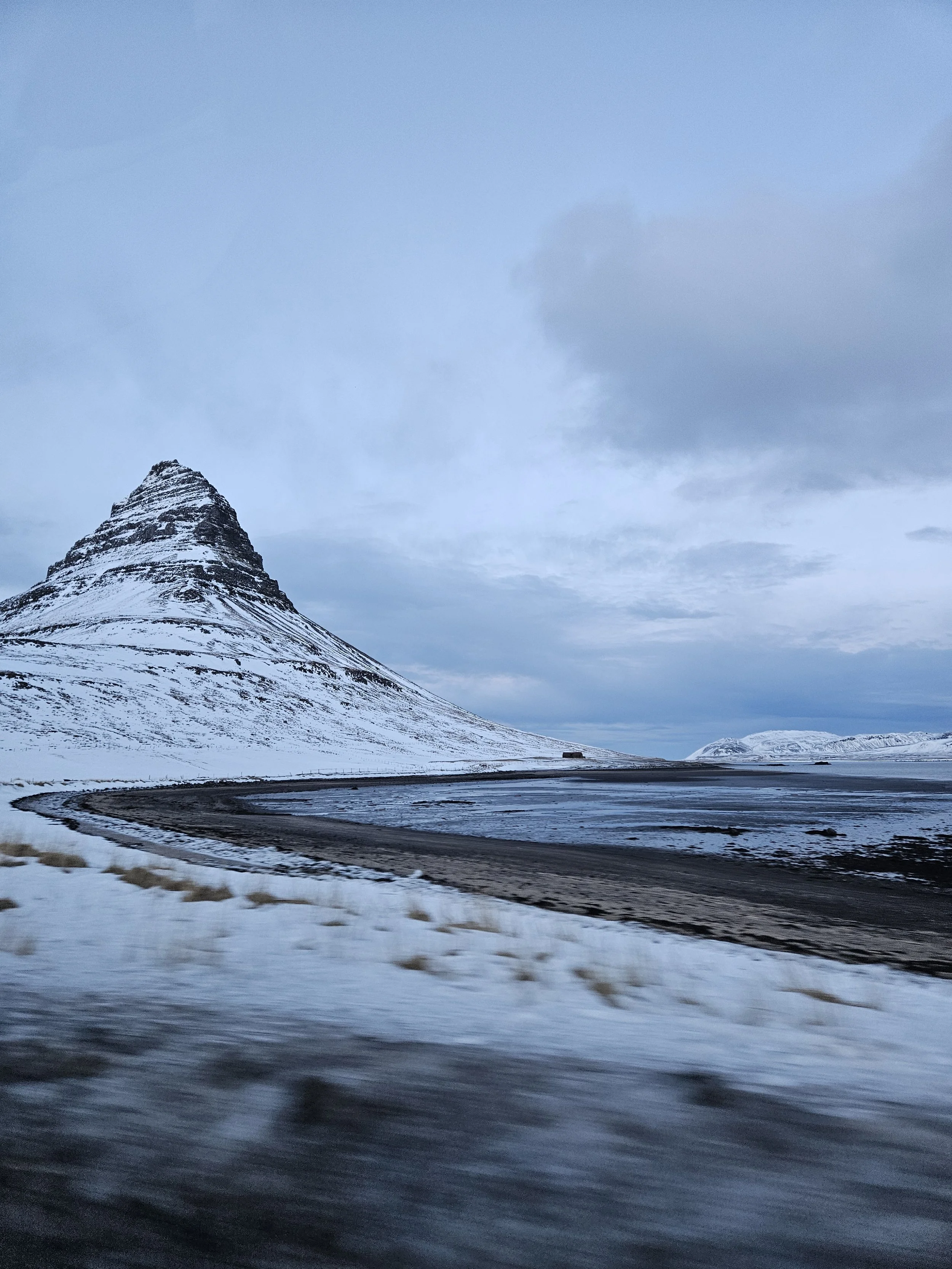 A snow-capped mountain with a conical shape, possibly a volcano, under a cloudy sky. A curved dark road runs through a snow-covered landscape in the foreground.