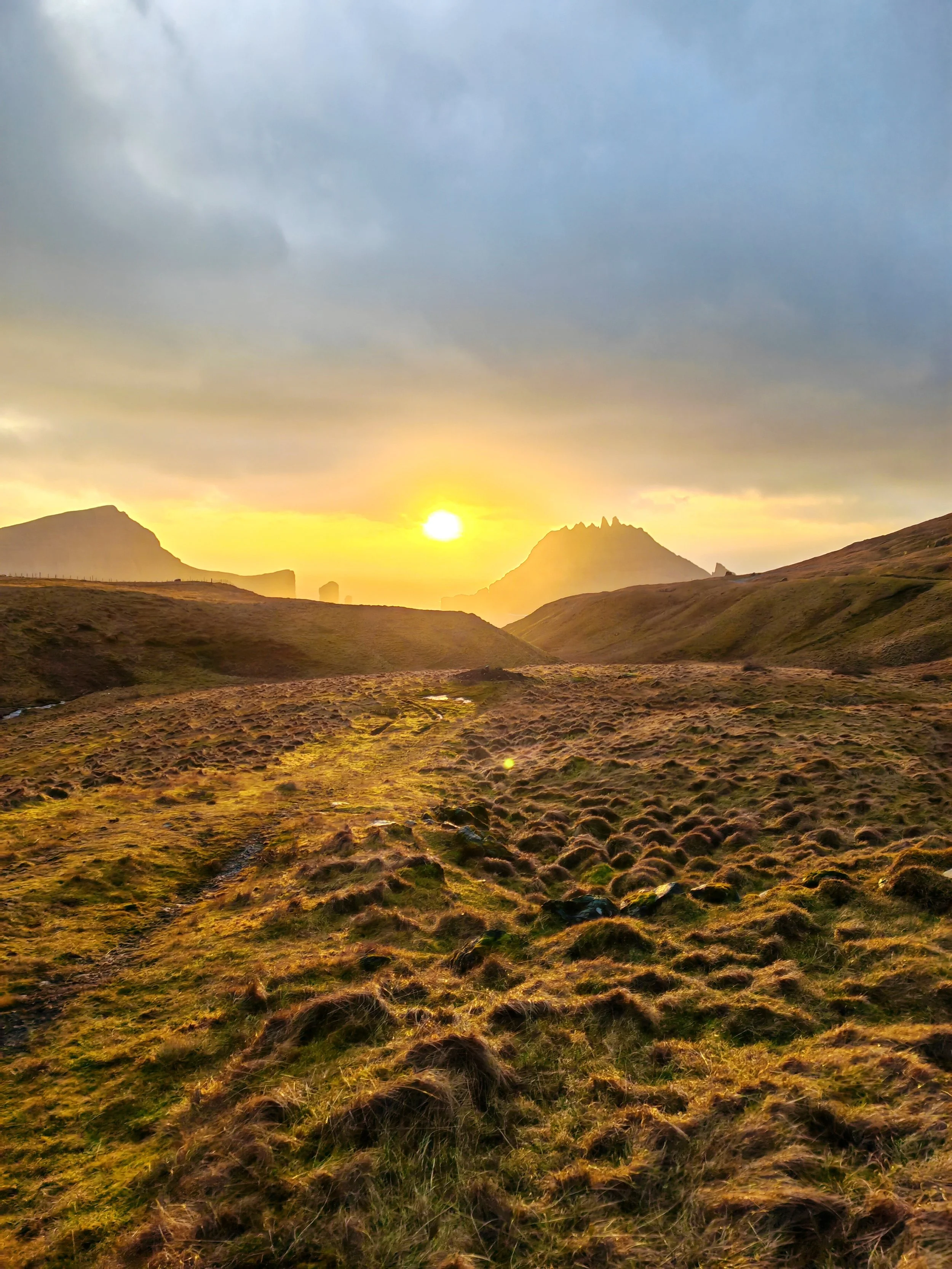 Sun setting behind mountains in a grassy landscape with cloudy sky