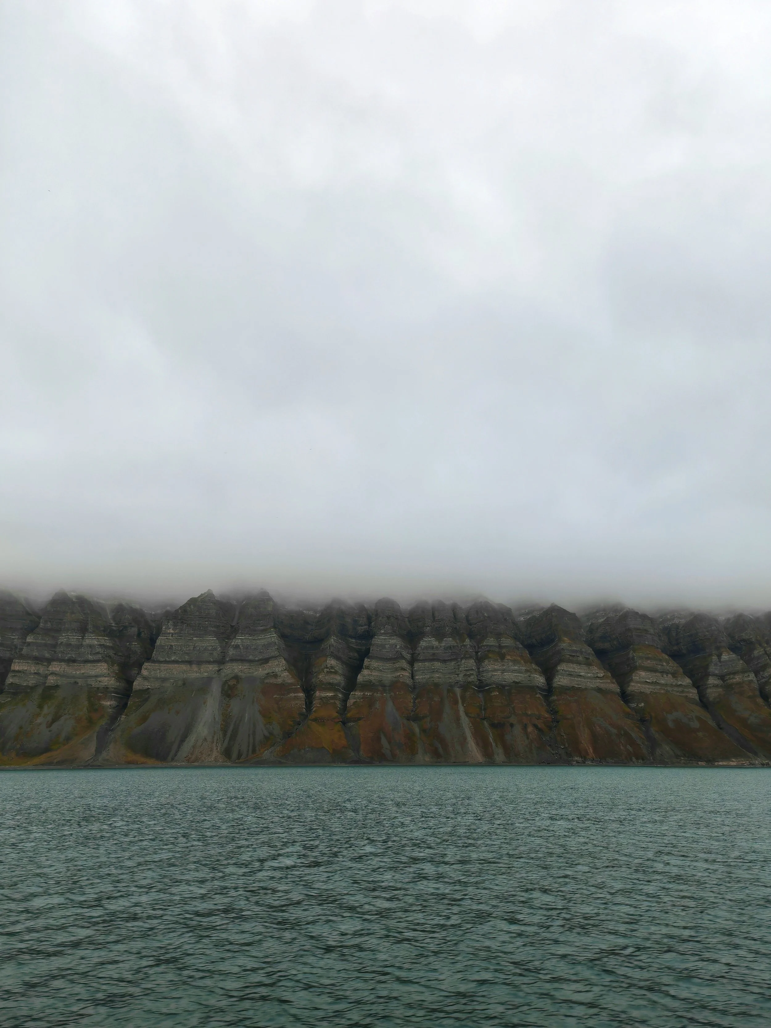View of a body of water in the foreground with a mountain range in the background. The mountain peaks are partially obscured by thick gray clouds.