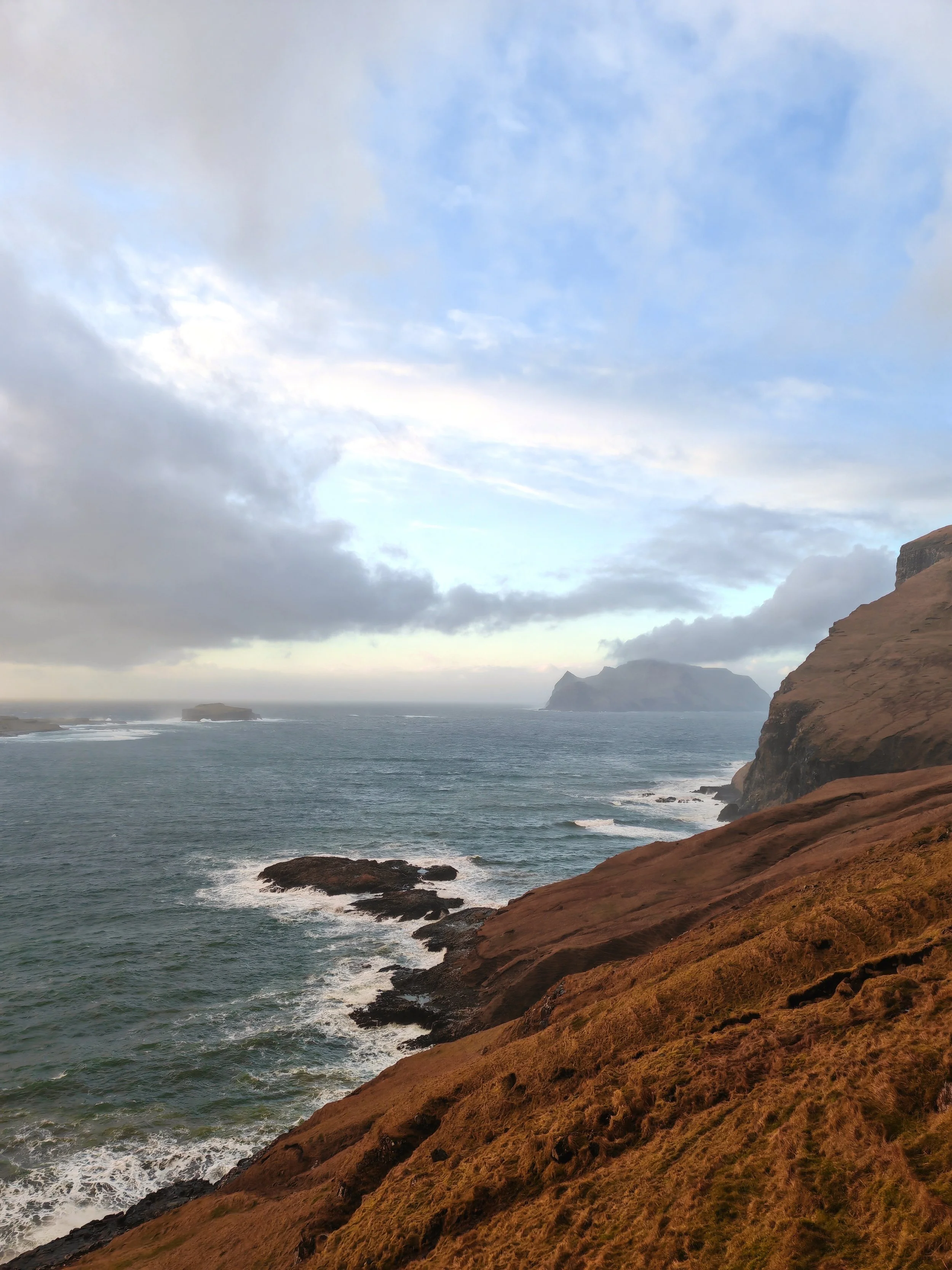 A coastal landscape with a rocky hillside in the foreground, the ocean in the middle distance, and cliffs and islands under a cloudy sky in the background.