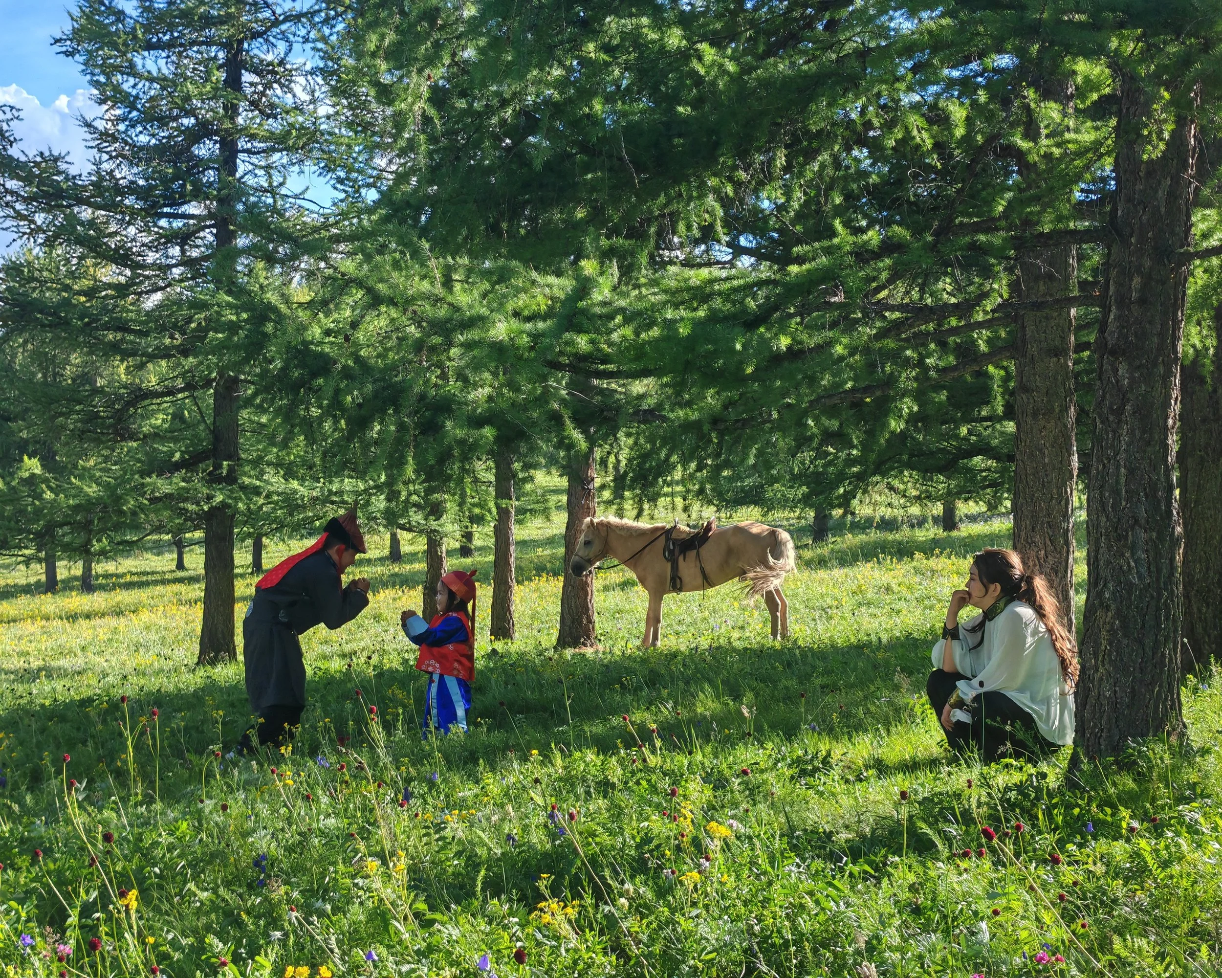 Children and an adult in a grassy field surrounded by tall trees with green leaves, some horses are present, and the children appear to be holding or interacting with flowers.