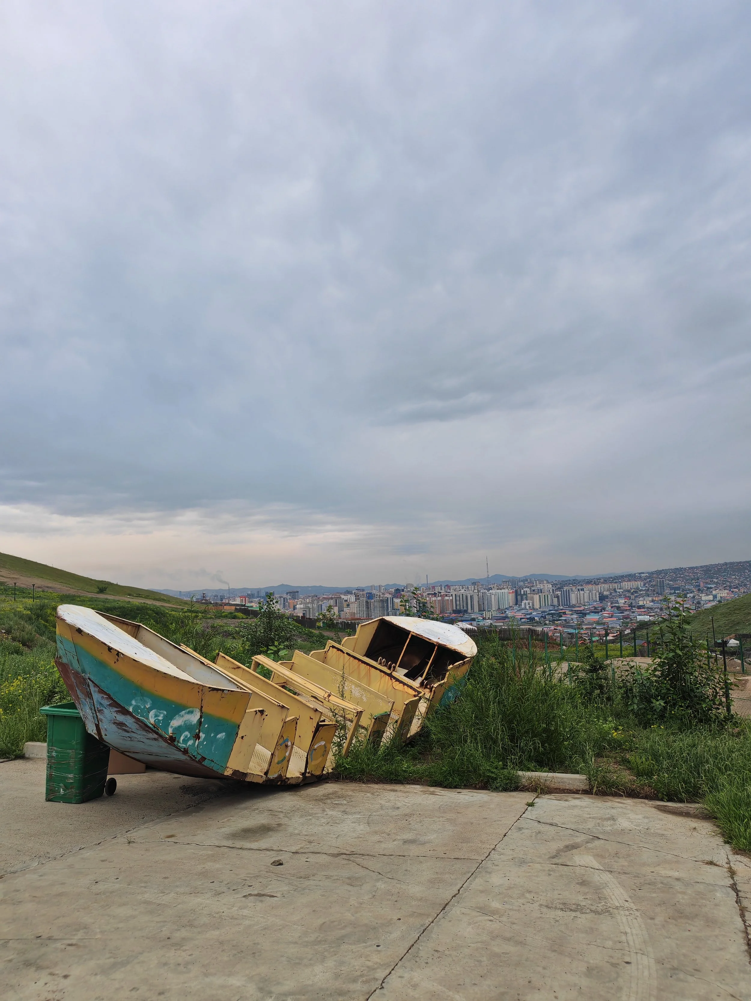 Empty, rusty boat leaning on a green trash bin on a concrete surface with a grassy hillside and city skyline in the background under cloudy sky.