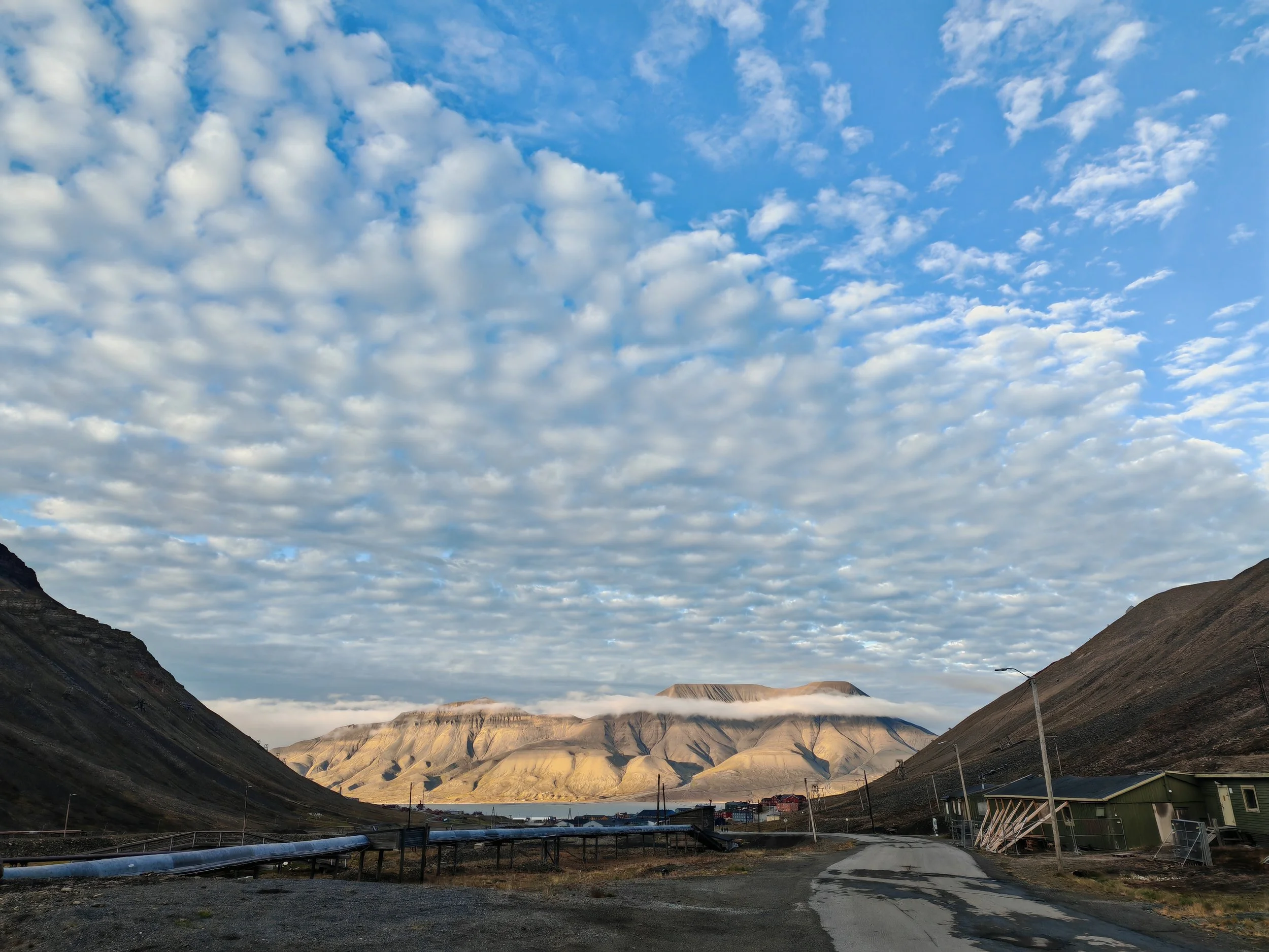 A mountain valley under a partly cloudy sky with hills and mountains in the background, and some buildings and infrastructure in the foreground.