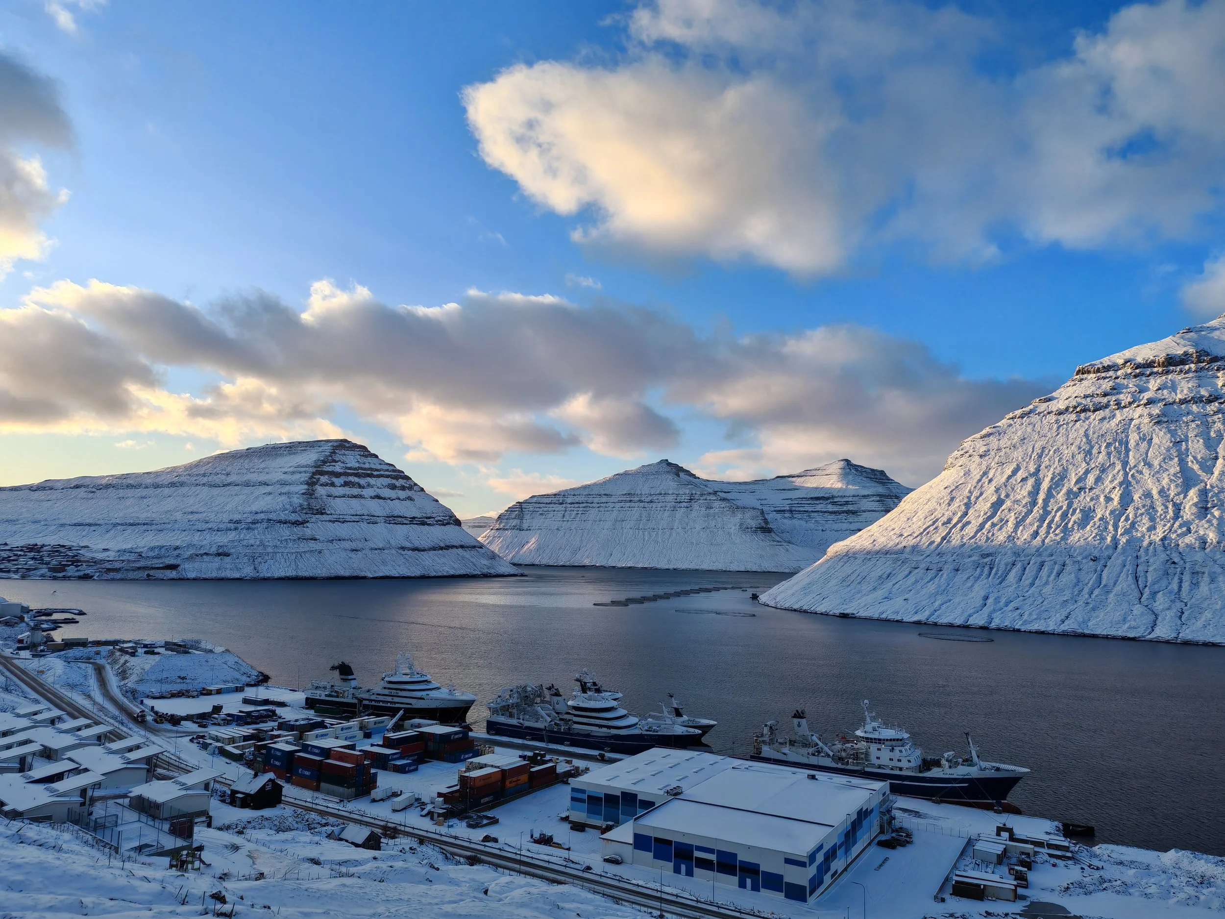 Snow-covered mountains overlooking a harbor with yachts and a small town with industrial buildings.