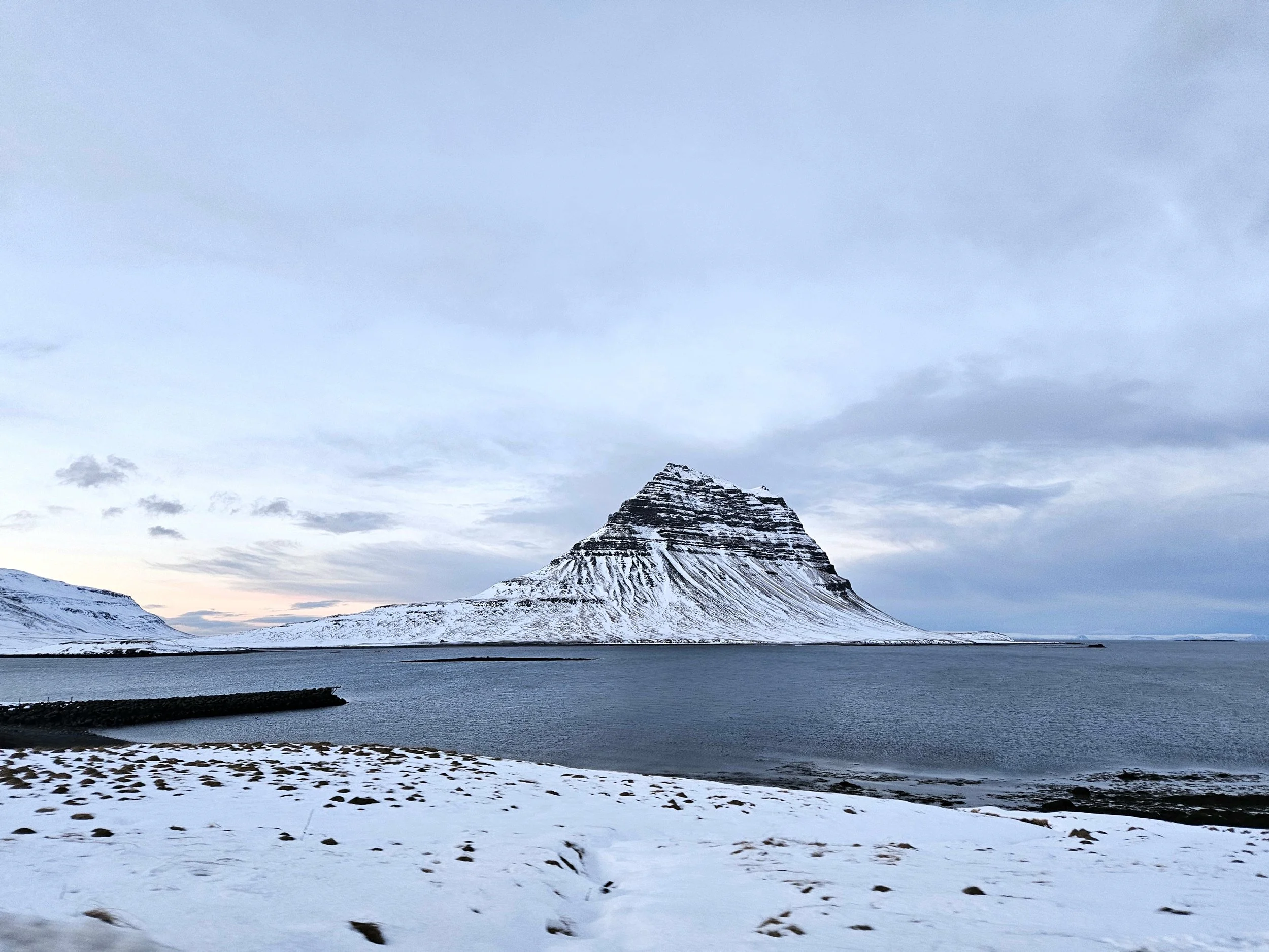 Snow-covered landscape with a mountain in the distance, a body of water, and a cloudy sky.