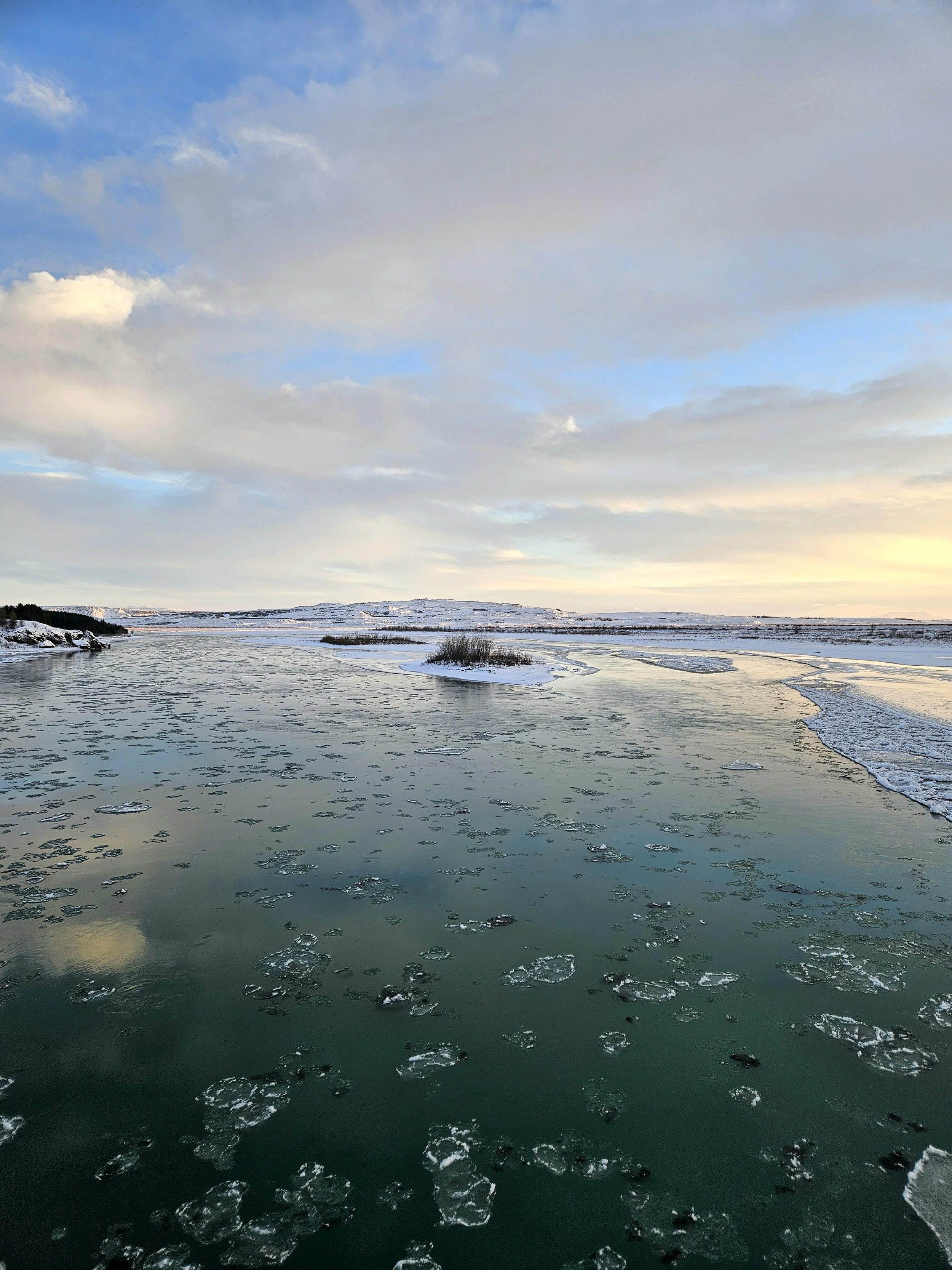 A frozen river with patches of ice floating on the surface, surrounded by snow-covered land under a partly cloudy sky.
