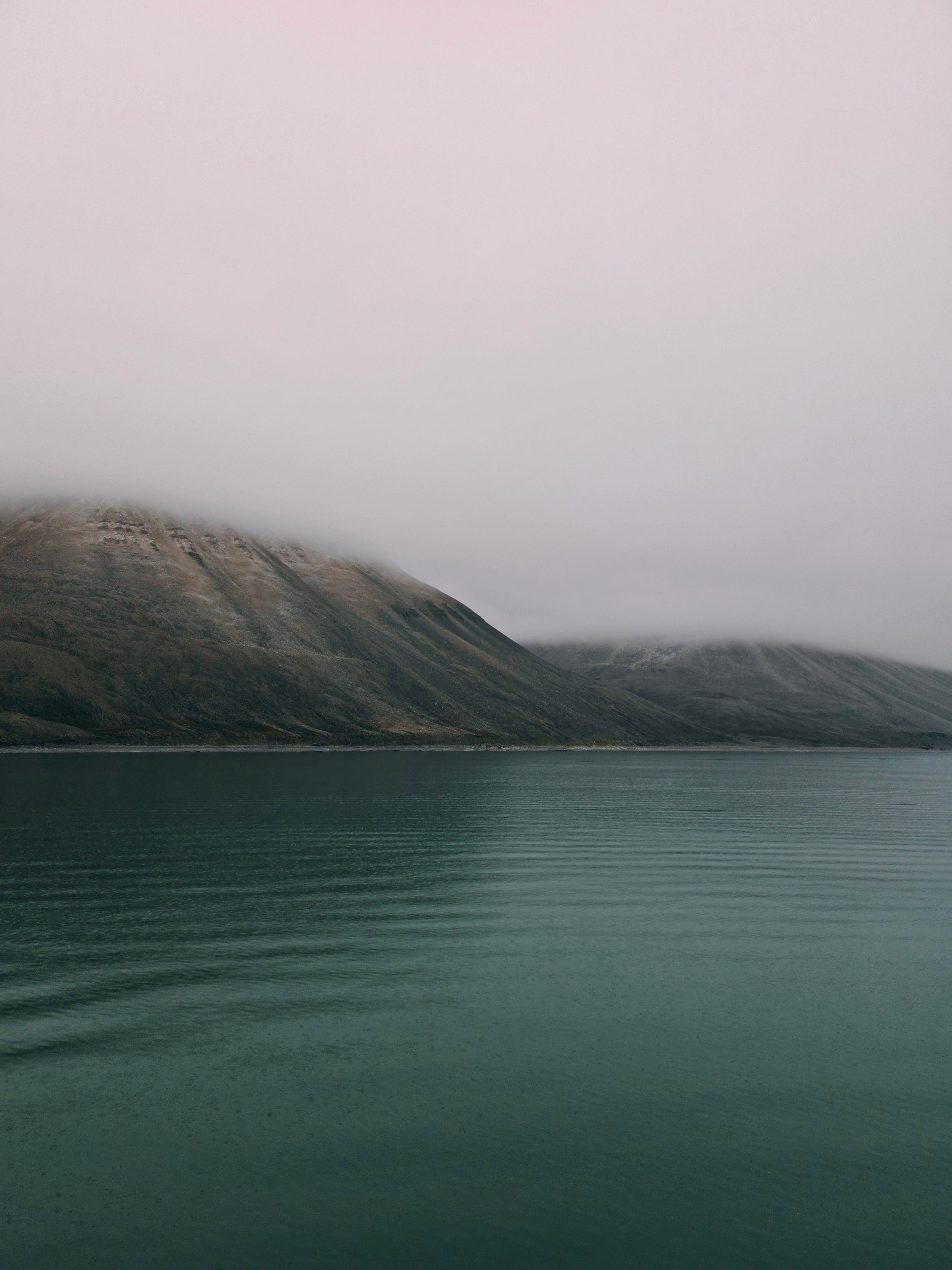 A calm body of water with mountains in the background, partially covered by fog and low clouds.
