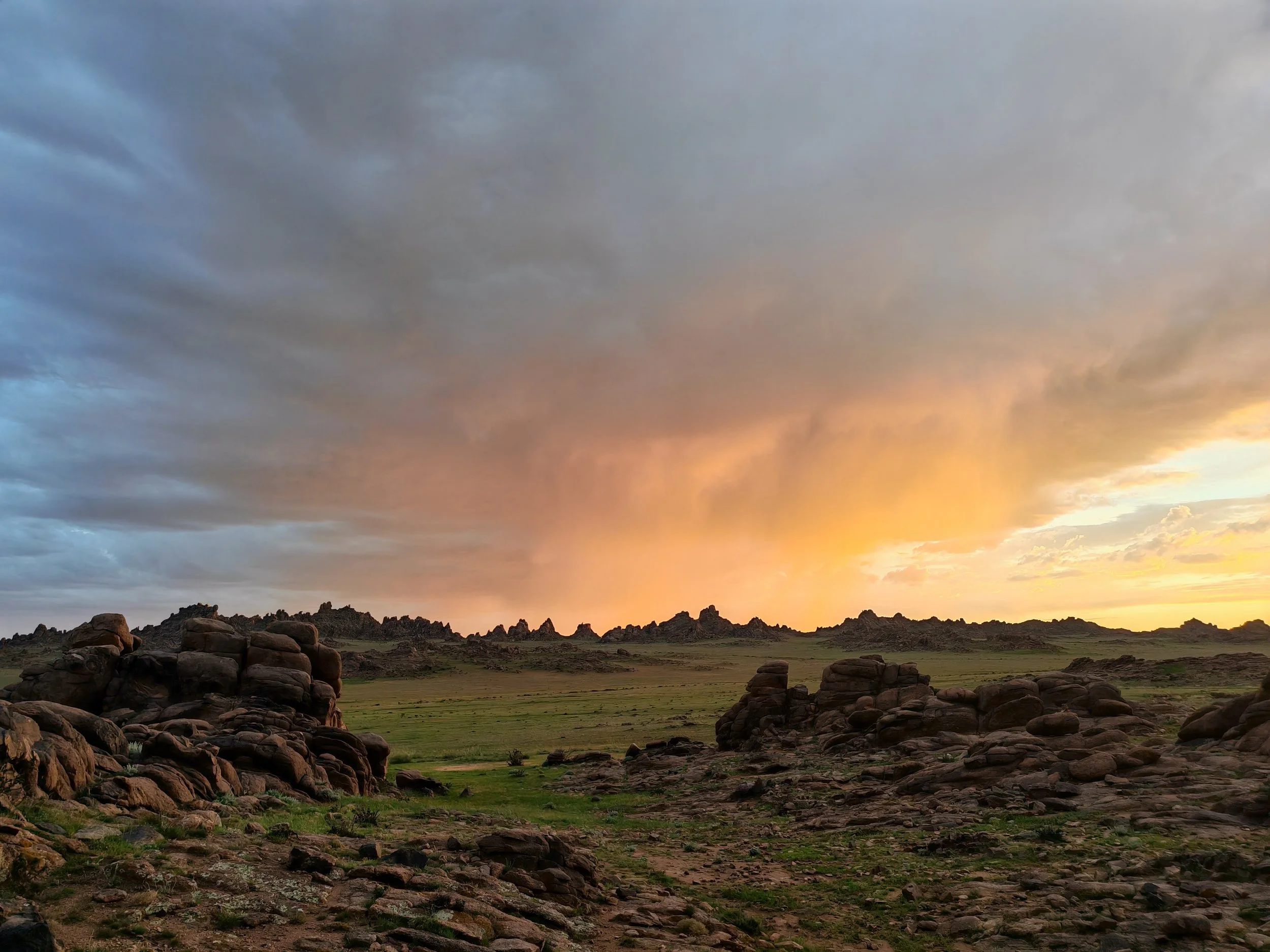 A landscape scene at sunset with dark clouds in the sky, rocky formations across the field, and a mixture of green grass and dry terrain in the foreground.