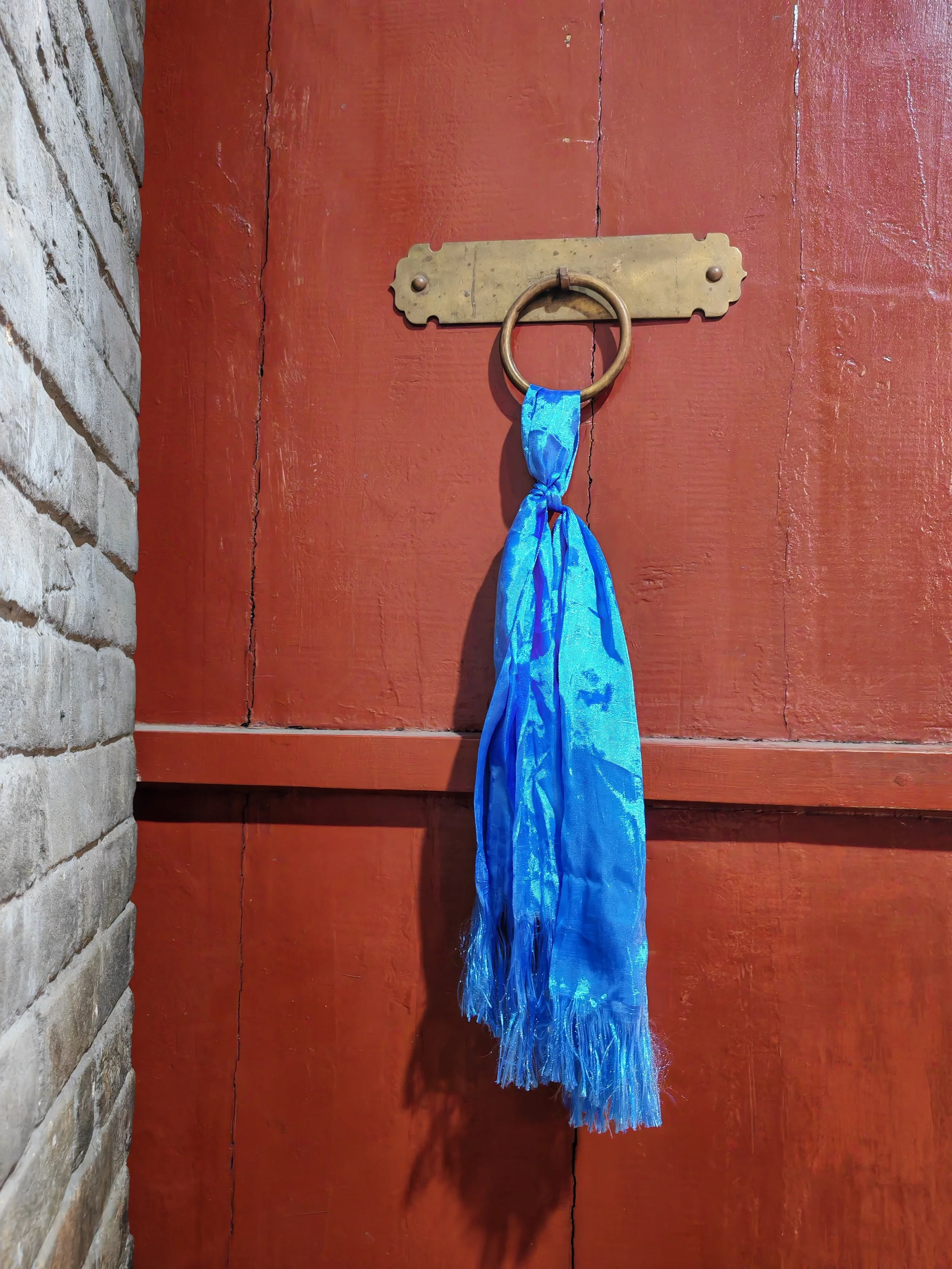 A brass door knocker with a blue cloth hanging from it, mounted on a red wooden door with visible wood grain and vertical panels.
