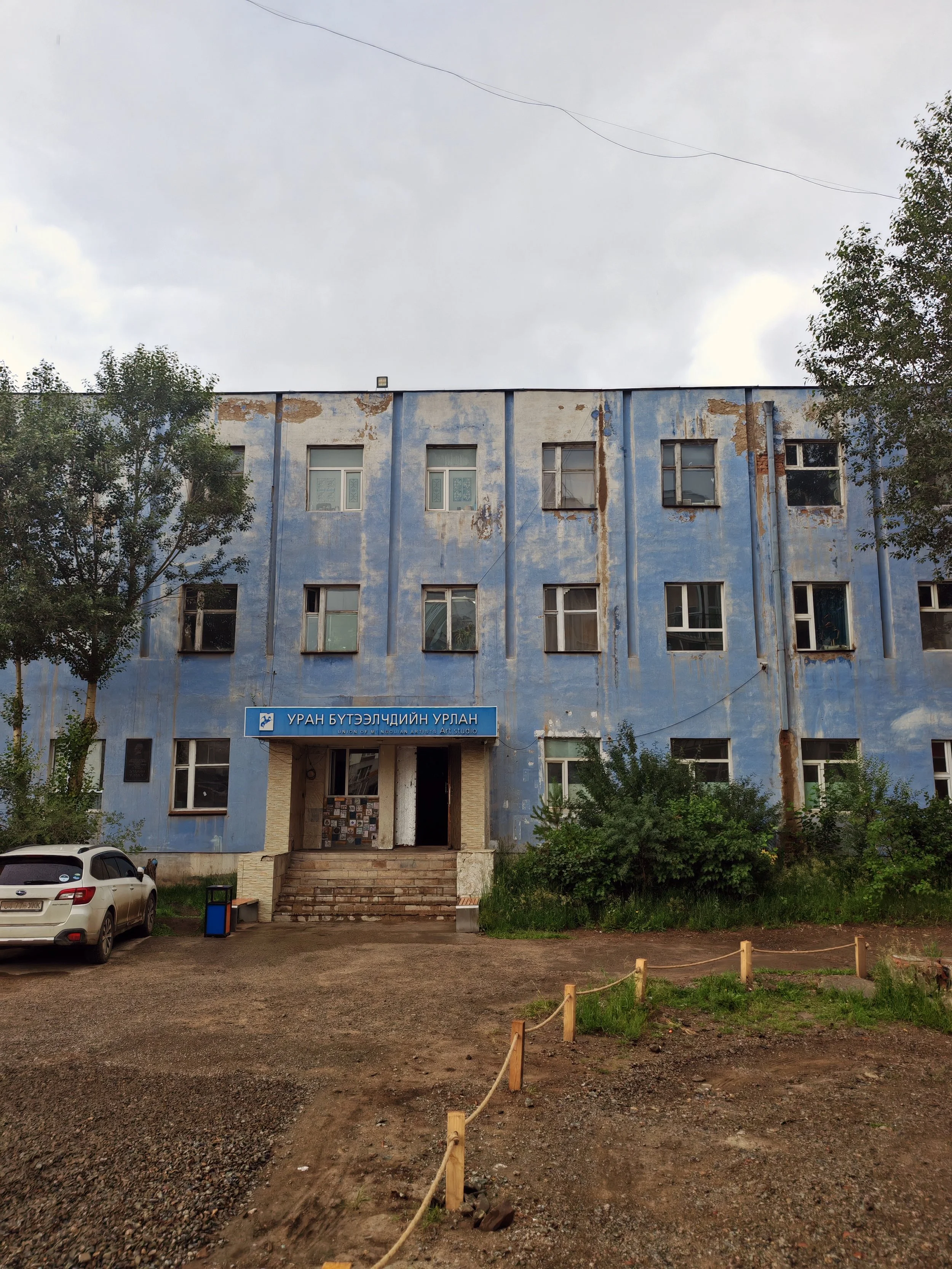 A blue, deteriorating building with peeling paint, multiple windows, and stairs leading to the entrance with a sign in Mongolian. Two cars are parked in front, and the area around has some trees, small plants, and a dirt lot with a makeshift wooden f