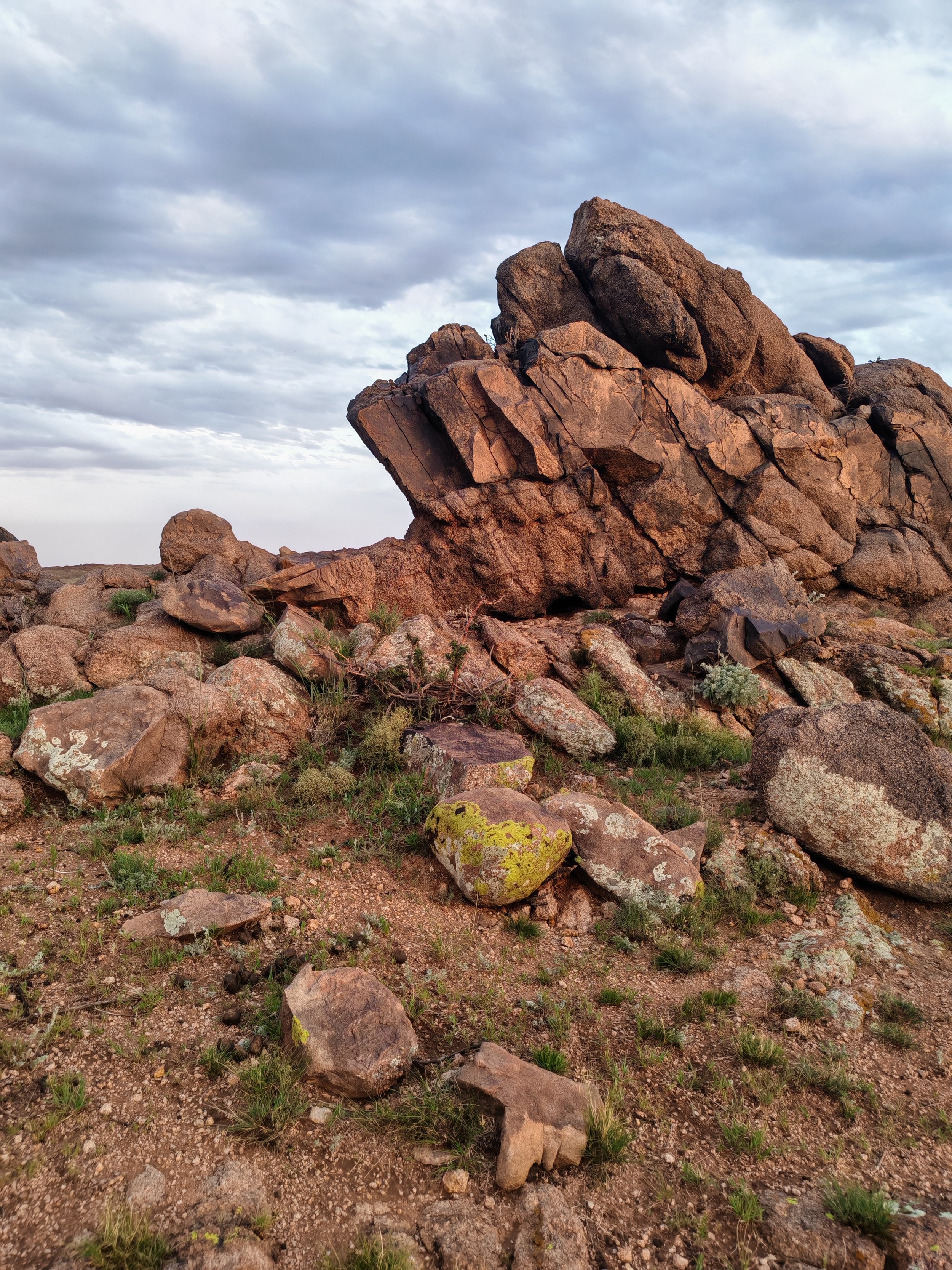A landscape with large reddish-brown rocks and boulders scattered across a dry, sparsely vegetated terrain, under a partly cloudy sky.