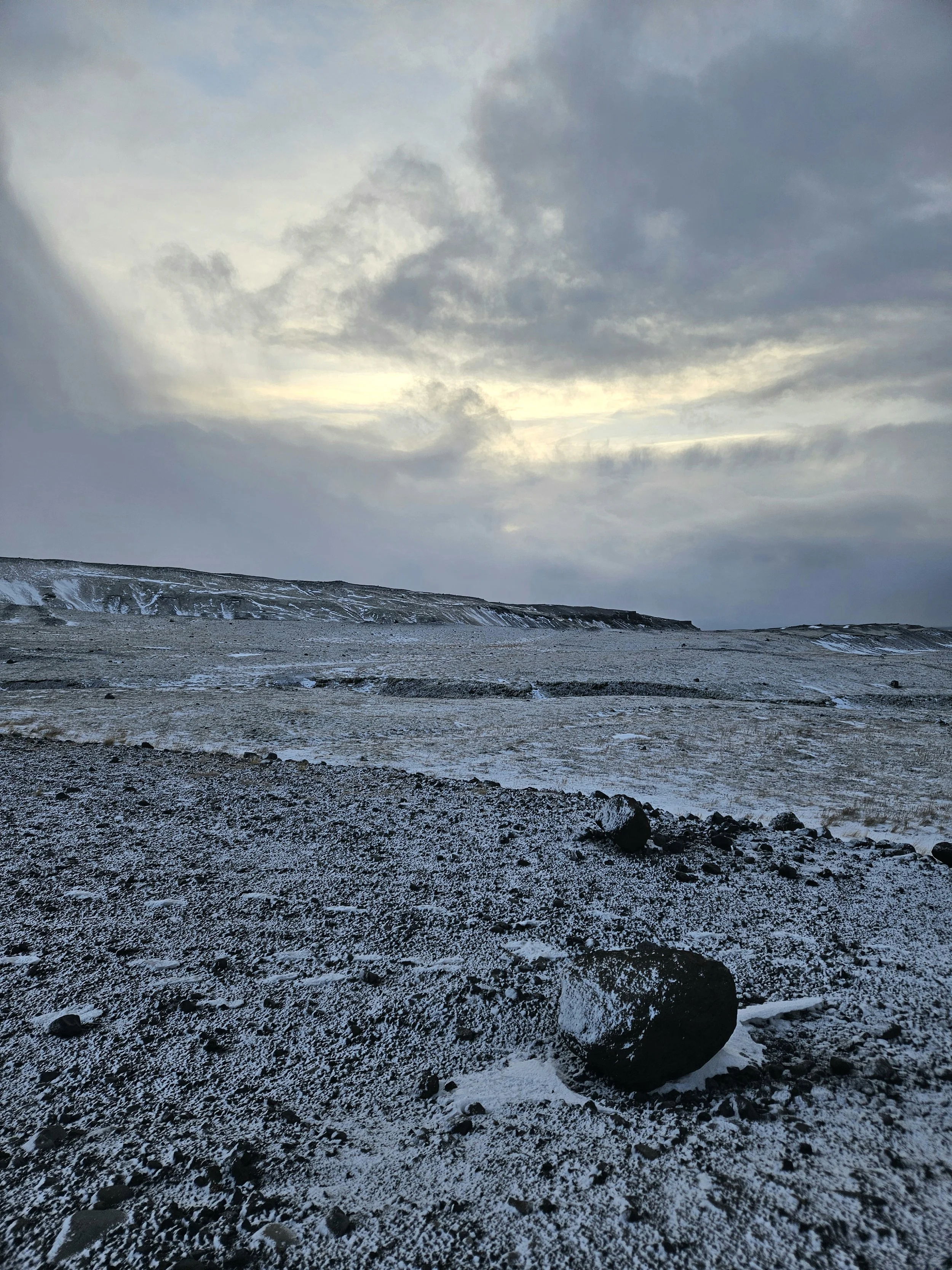 A rugged, snow-dusted landscape with a dark, rocky foreground and a barren, snow-covered plains stretching to distant cliffs under a cloudy sky.