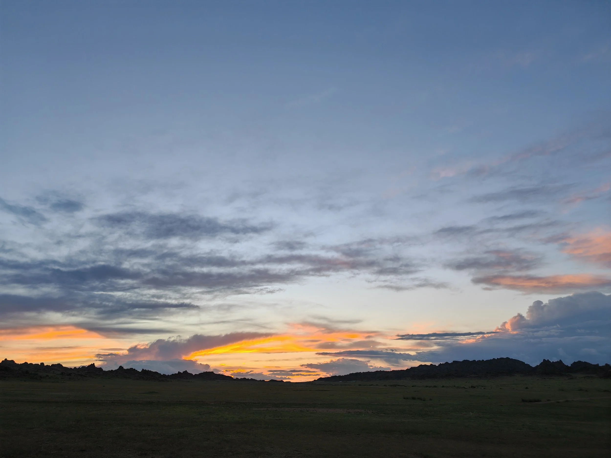 A landscape at sunset with a dark, rocky horizon, and a partly cloudy sky featuring shades of blue, pink, and orange.