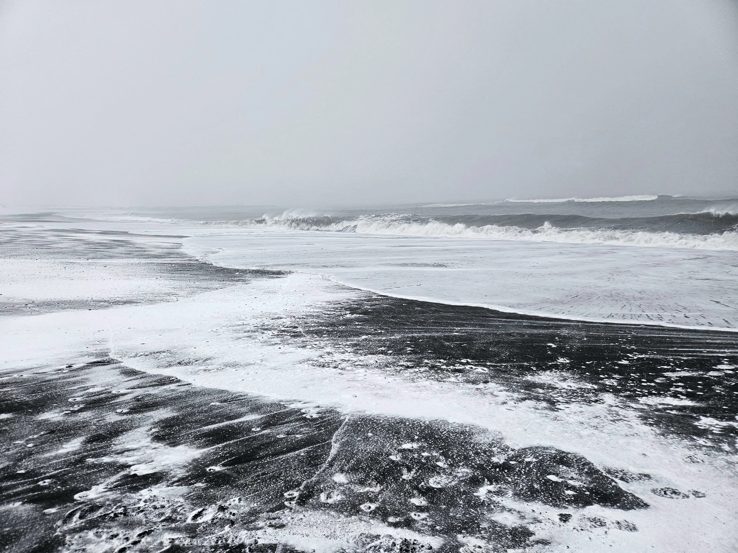 Overcast day at a beach with waves crashing onto the shore, a mix of dark sand and light snow or foam on the beach, with a hazy, gray sky.
